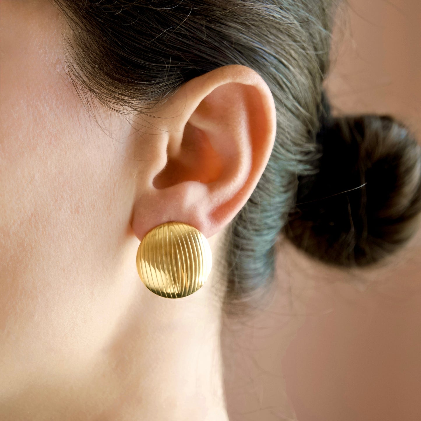 A close-up of a person's ear wearing a textured, round gold stud earring against a soft beige background.