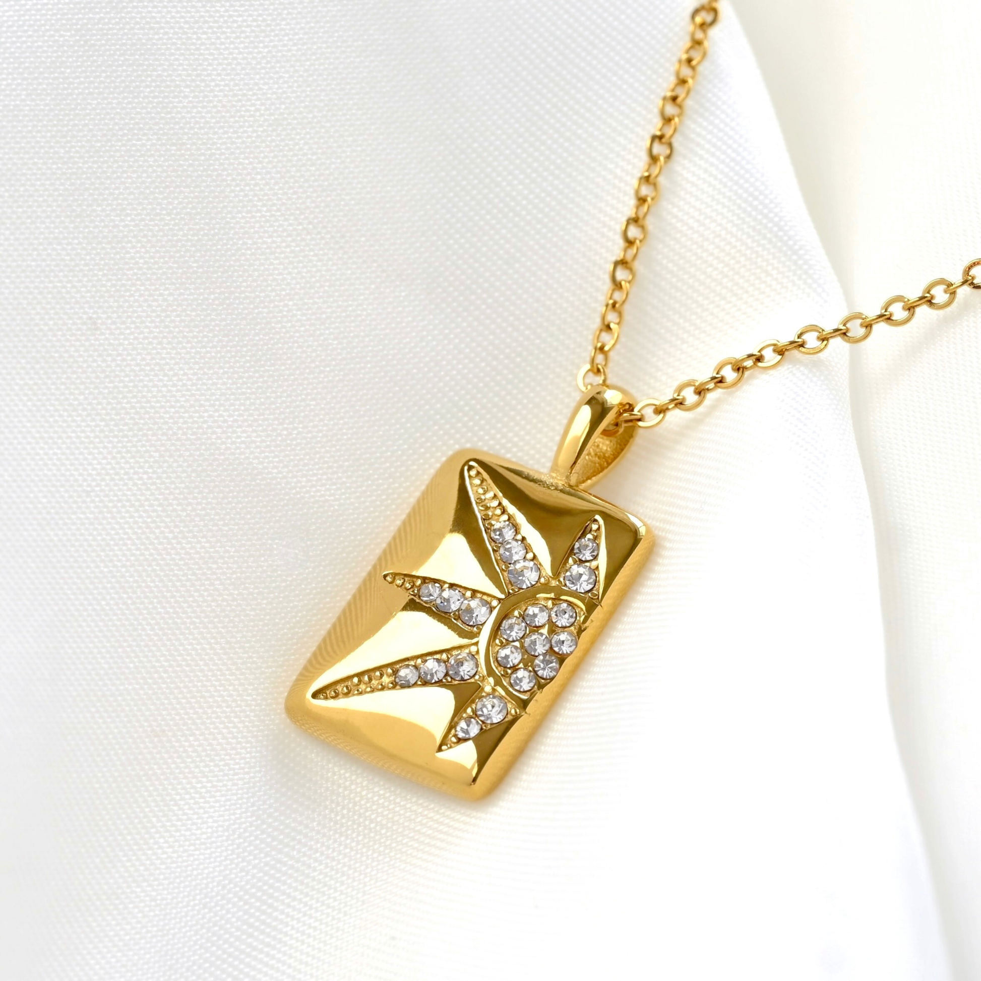 A gold rectangular pendant necklace featuring a starburst design set with small clear crystals, displayed on a draped white fabric background.