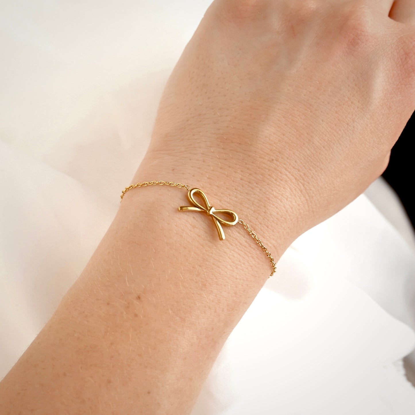 A delicate gold chain bracelet with a small bow charm, shown worn on a person's wrist against a soft white background.