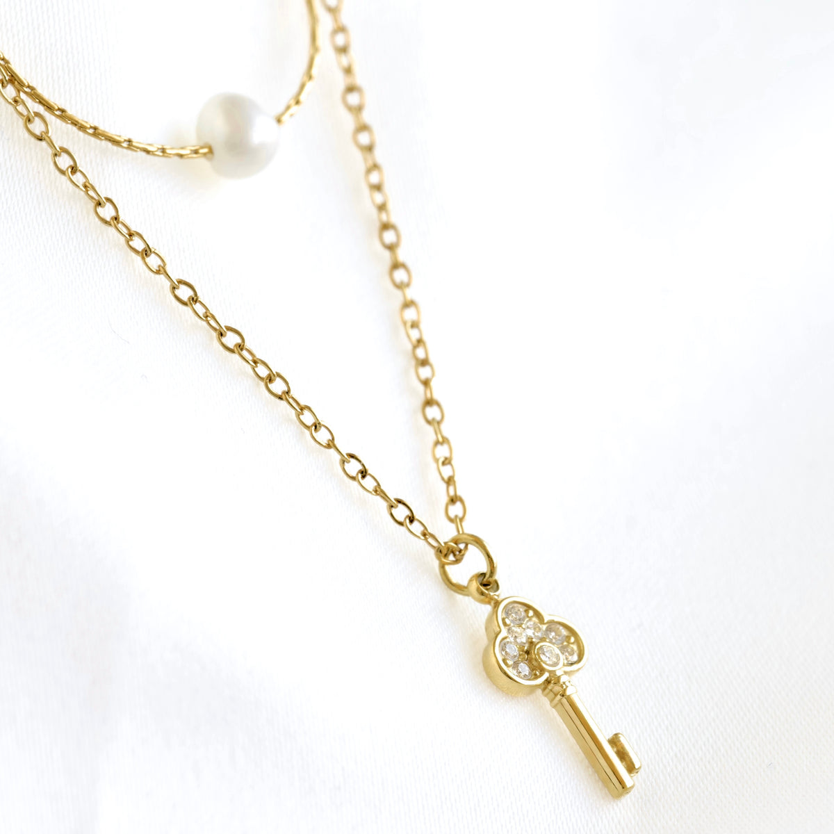 A gold key pendant necklace with clear crystal accents, displayed on a delicate gold chain alongside a pearl bead necklace on a white fabric background.