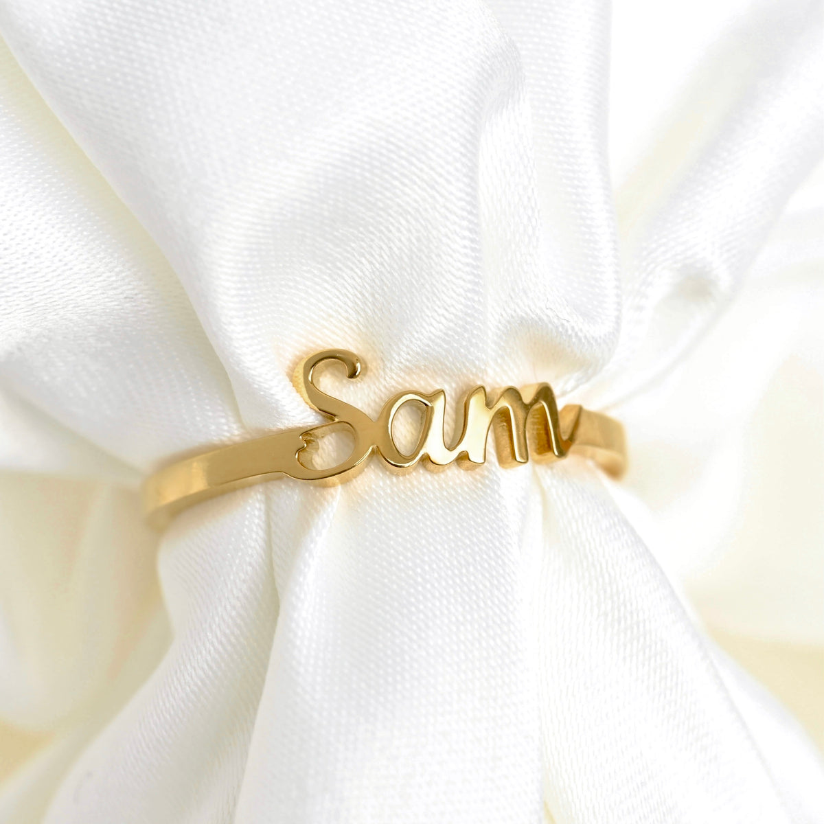 A gold name ring engraved with the name 'Sam' in cursive, displayed on a white satin fabric.
