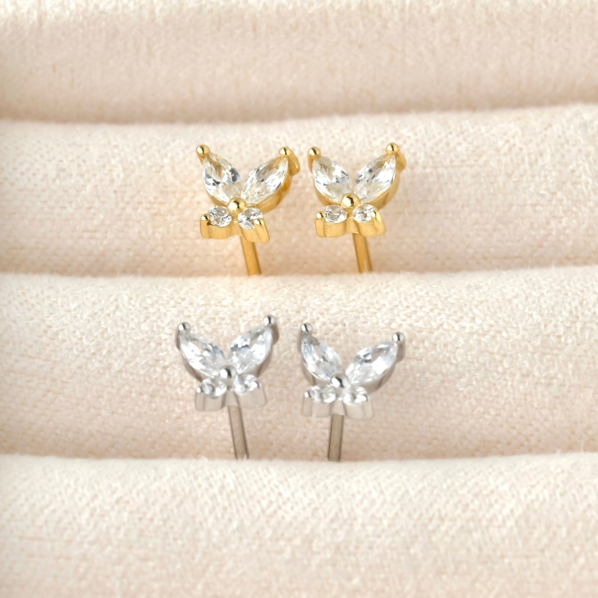 Butterfly-shaped CZ earrings in gold and silver on a beige surface