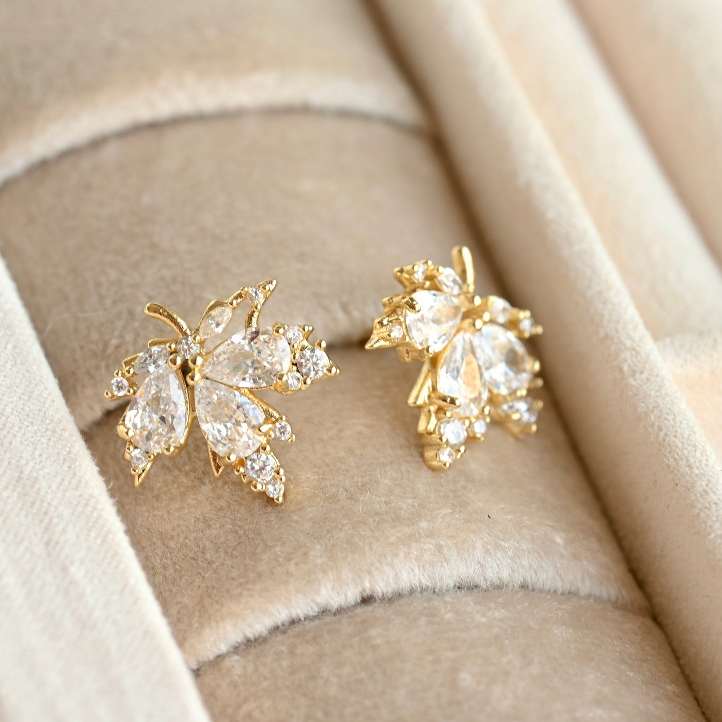 A pair of gold maple leaf-shaped stud earrings set with clear marquise and round CZ stones, displayed on a beige velvet jewelry box.