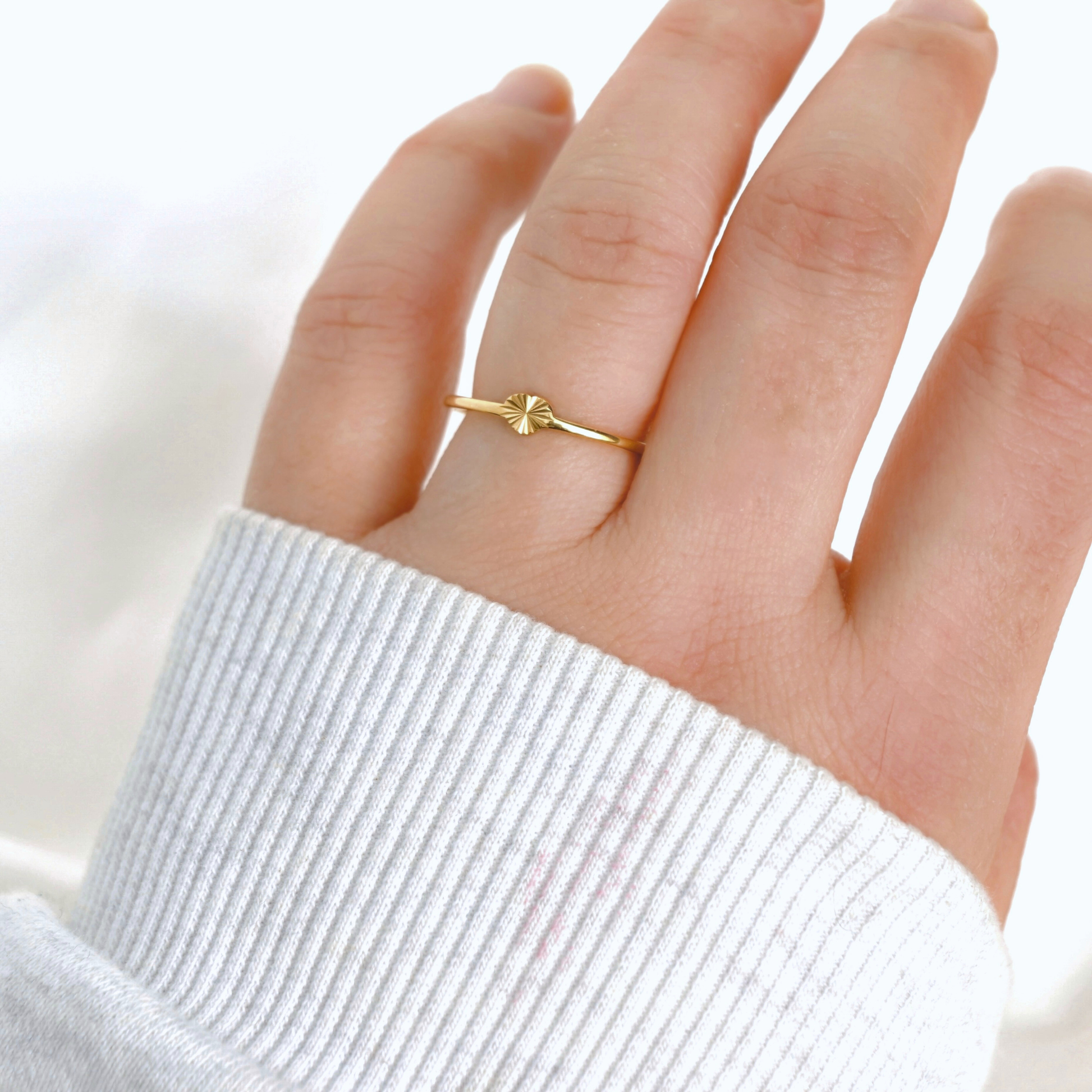 A delicate gold ring with a small starburst or sunburst motif on the front, worn on a person's ring finger. The hand is covered by a white ribbed knit sweater sleeve, set against a bright, soft-focus white background.