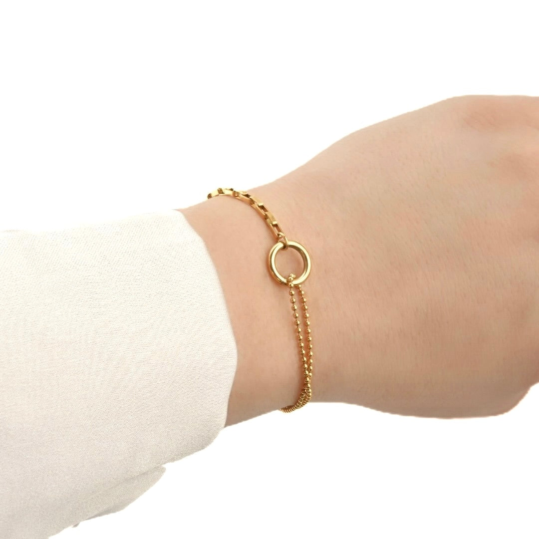 Gold bracelet with a mix of small old paperclip chain and a two layered bold chain linked with a small thick circle in the front, worn on a wrist with a plain background