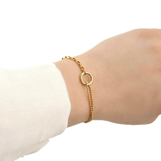 Gold bracelet with a mix of small old paperclip chain and a two layered bold chain linked with a small thick circle in the front, worn on a wrist with a plain background