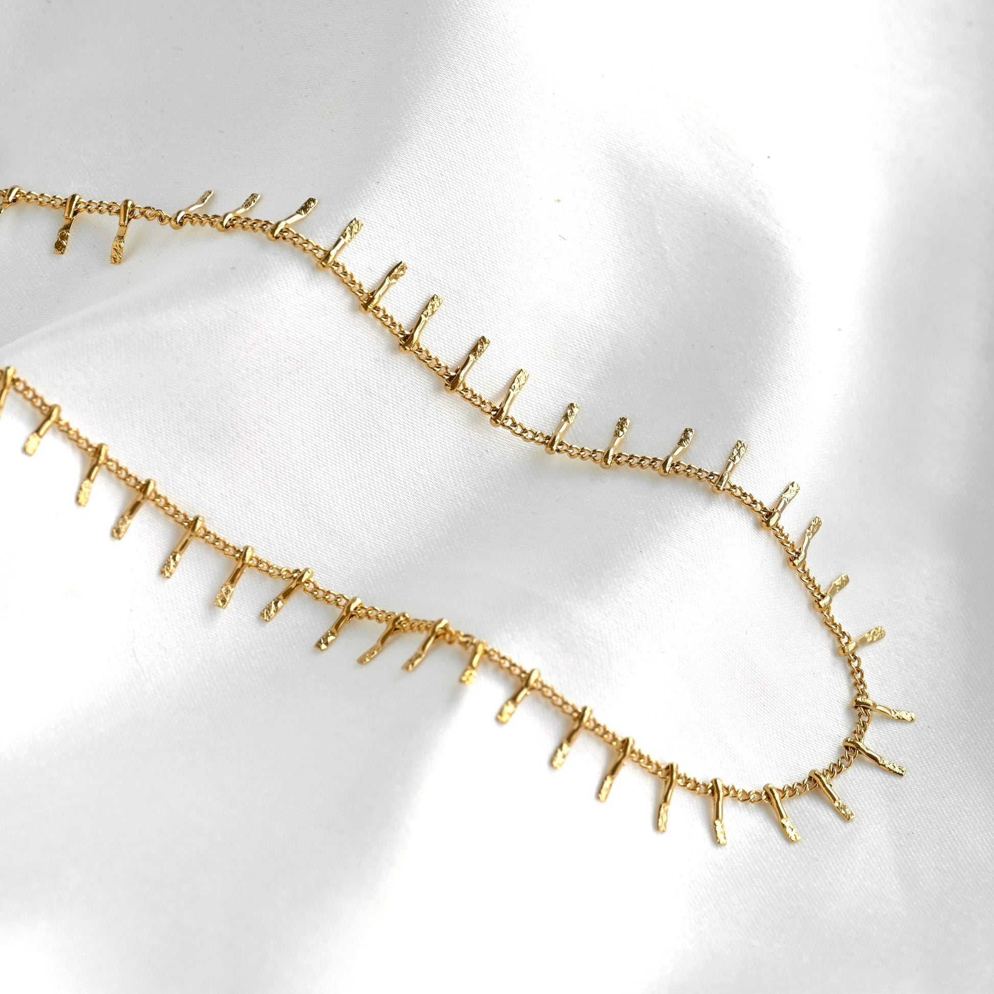 A gold chain necklace featuring small, evenly spaced textured spikes along the front, displayed on a soft white satin fabric background.