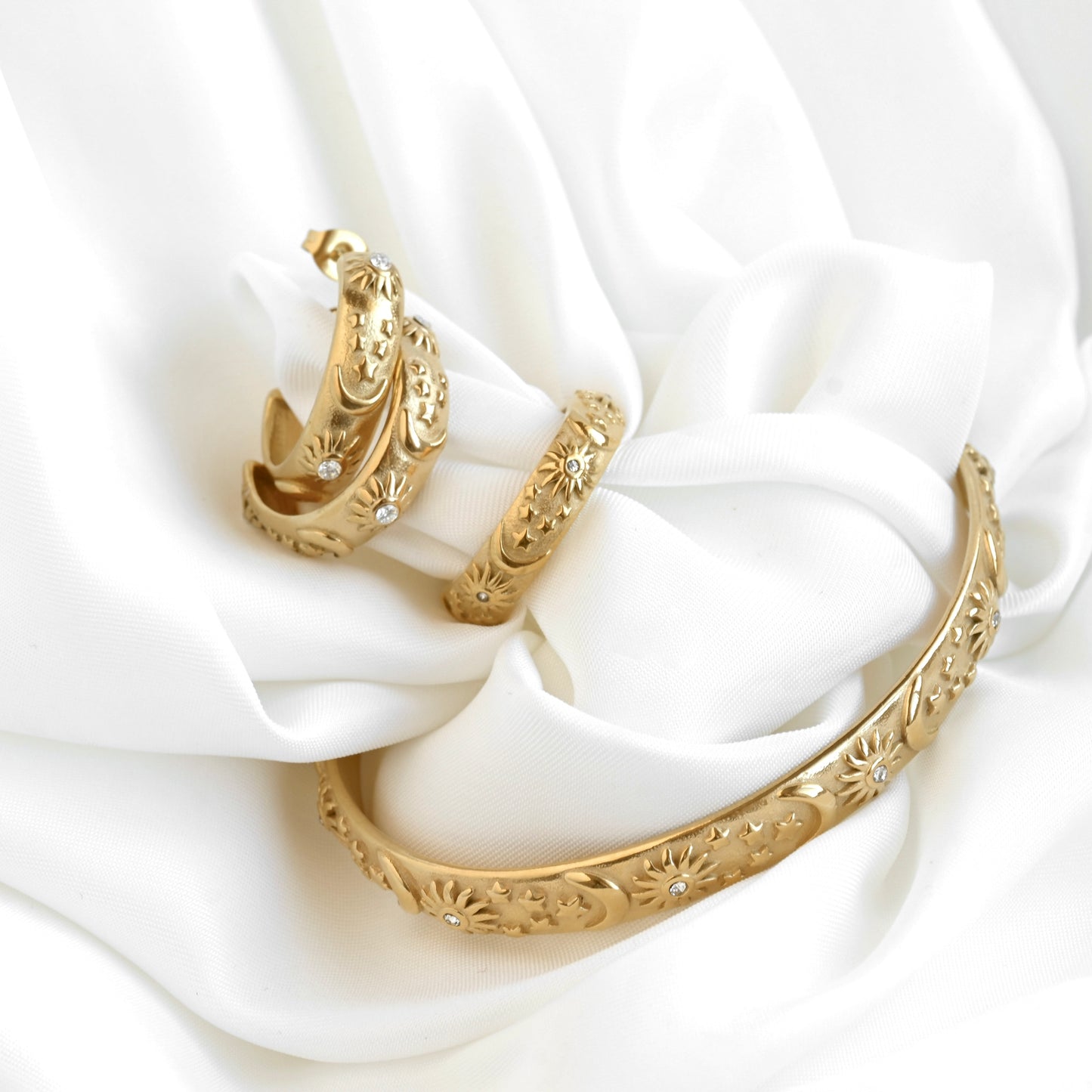 Gold jewelry set including a cuff bracelet, ring  and a pair of medium sized C hoops with celestial patterns on a white fabric background