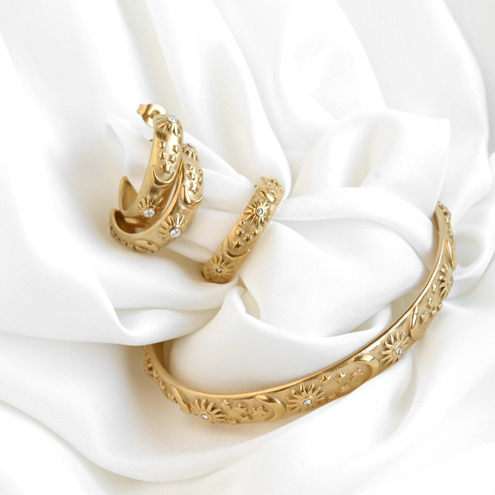 Gold jewelry set including a cuff bracelet, ring  and a pair of medium sized C hoops with celestial patterns on a white fabric background