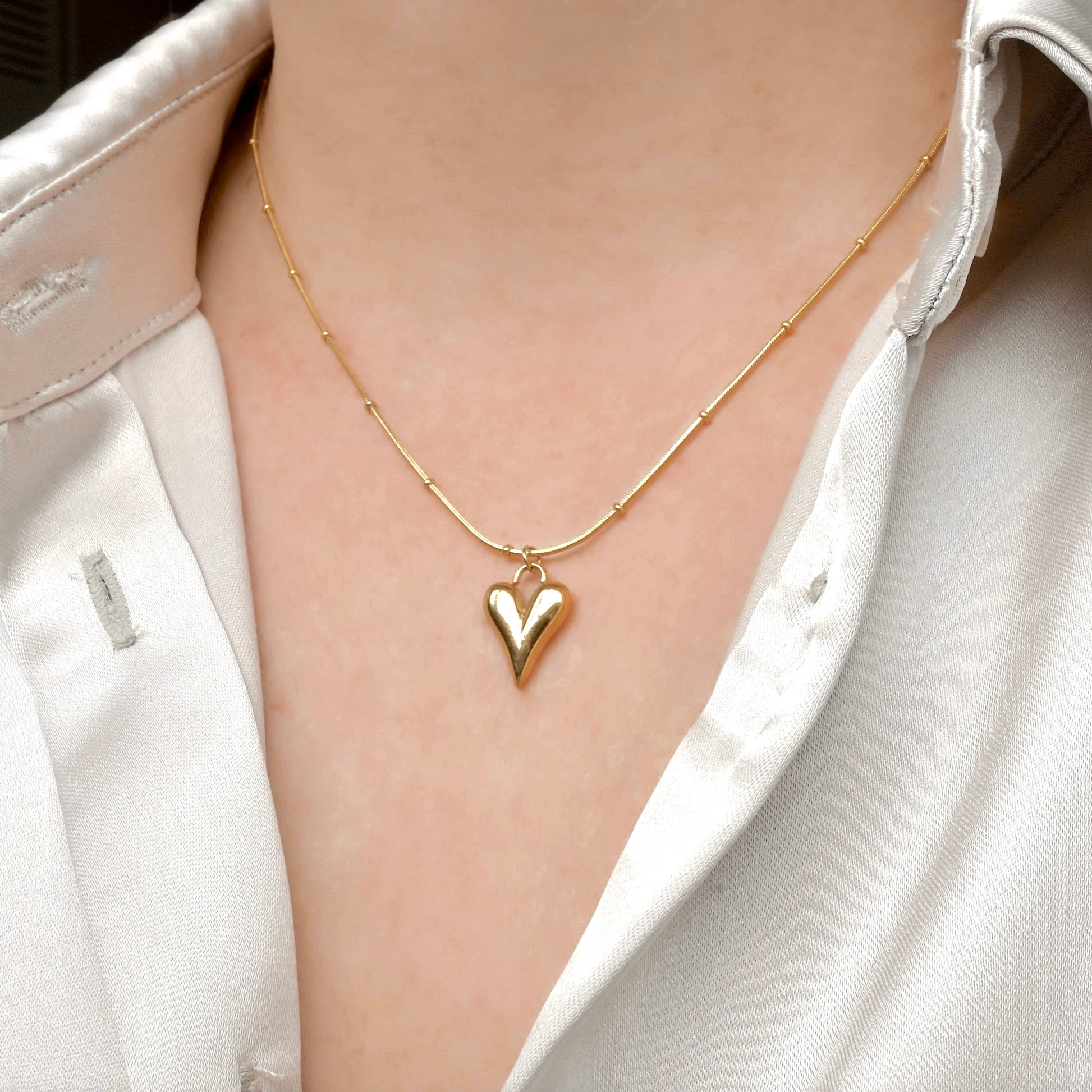 Angled view of gold puffy heart necklace worn by a person in a white shirt