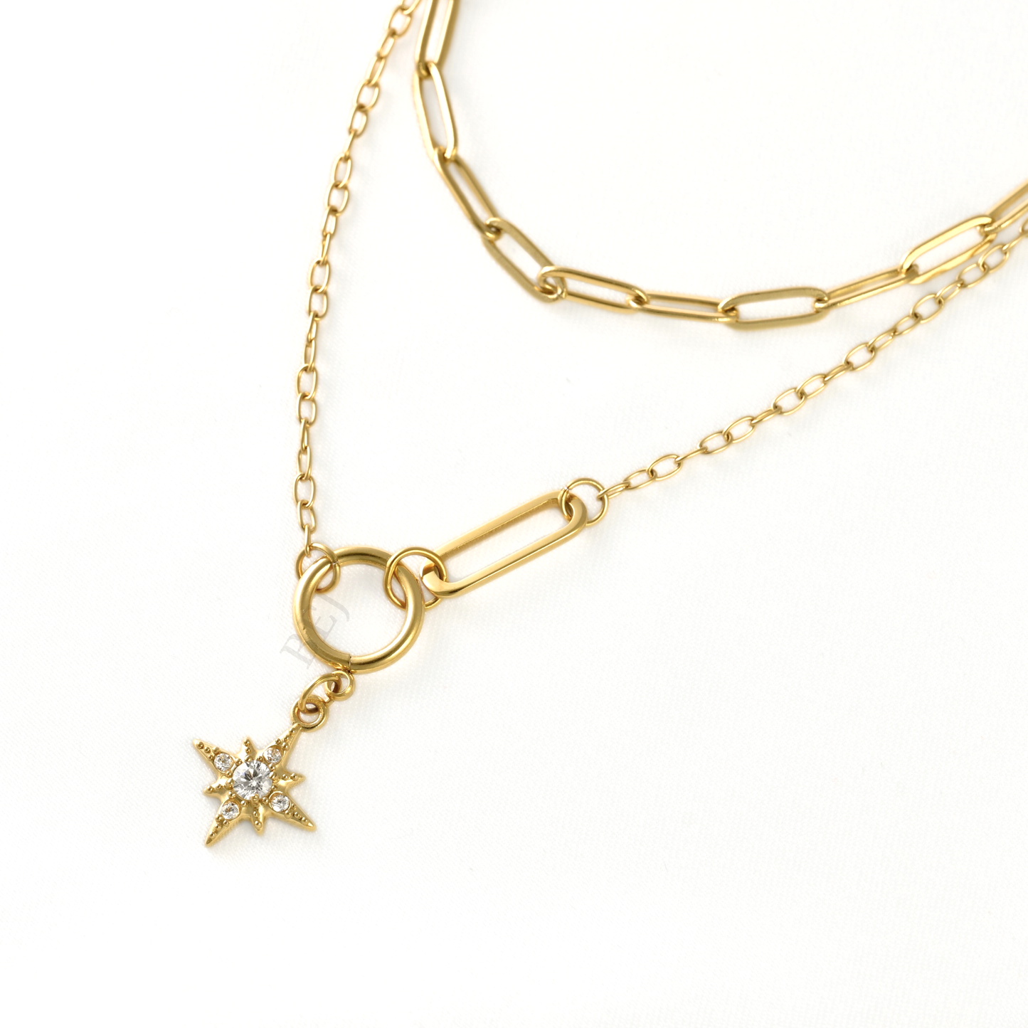A gold-toned layered necklace featuring a paperclip chain, a delicate cable chain, and a small starburst charm with a clear rhinestone, displayed on a clean white background.