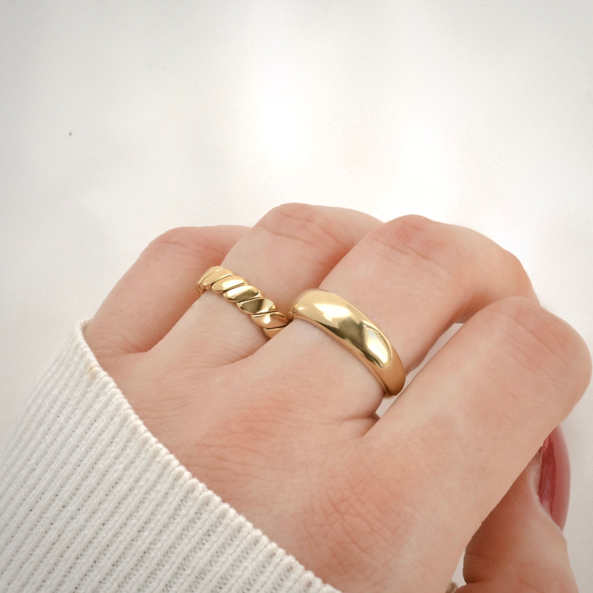 Twisted gold ring stacked with a chunky dome ring on a finger against a plain background