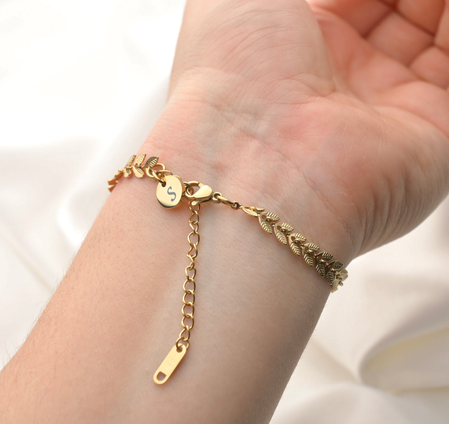 Gold chevron bracelet on a wrist with a small initial disc charm at the clasp on a plain white background