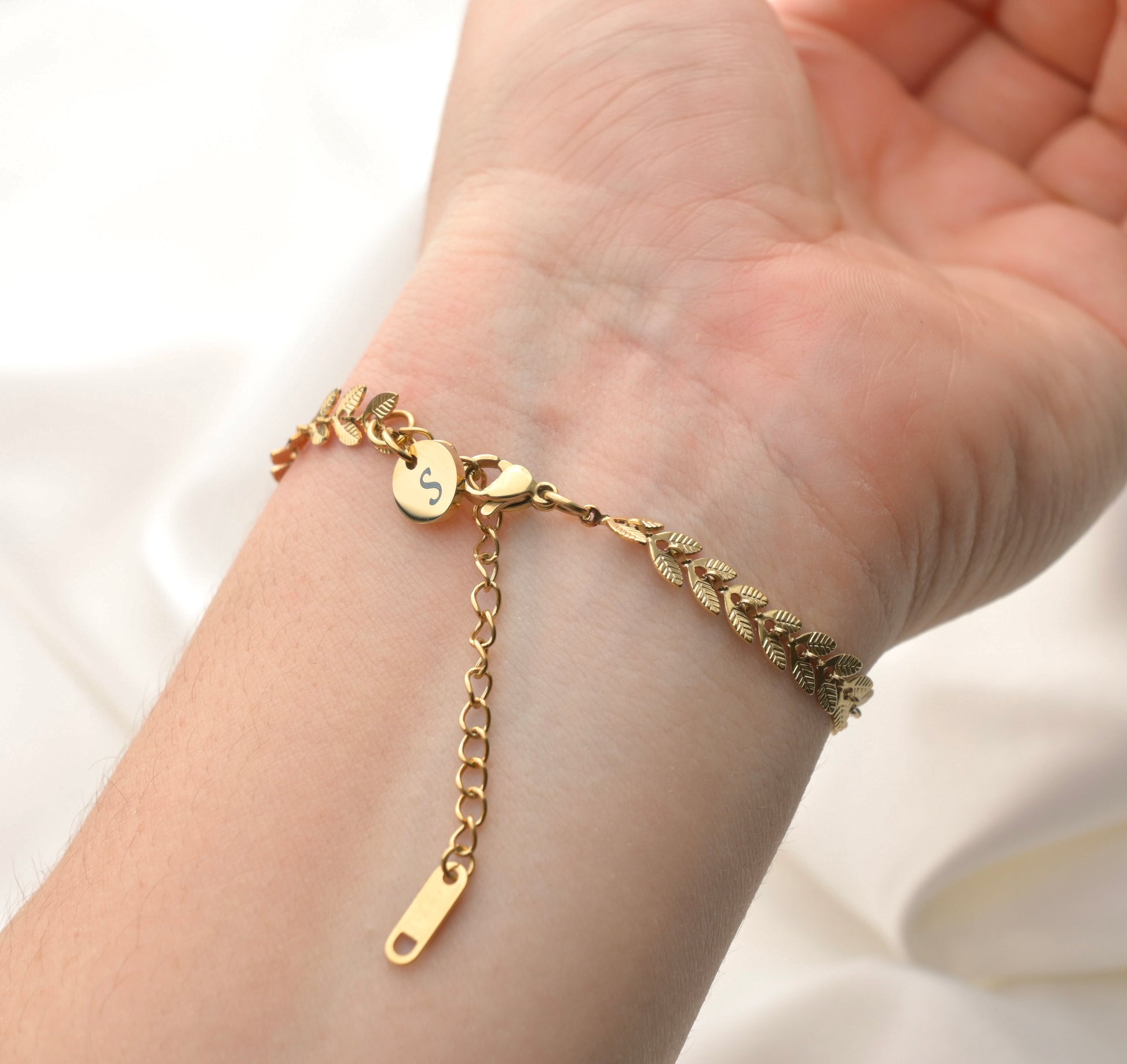 Gold chevron bracelet on a wrist with a small initial disc charm at the clasp on a plain white background