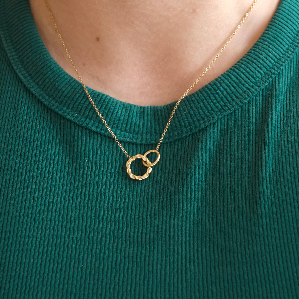 A close-up of a person wearing a delicate gold chain necklace with two interlocking textured rings pendant, styled over a dark green ribbed top.