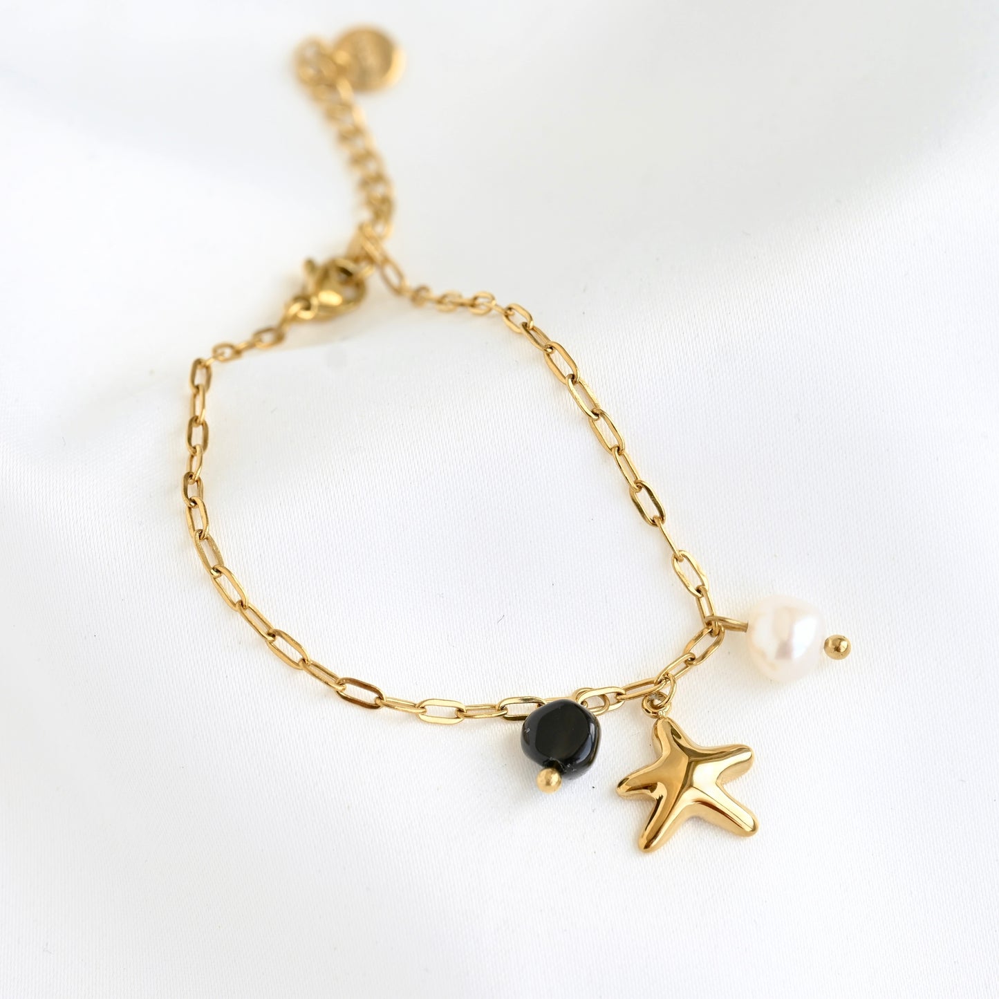 A delicate gold chain bracelet featuring a starfish charm, a small black stone, and a white natural pearl accent, displayed on a clean white background.