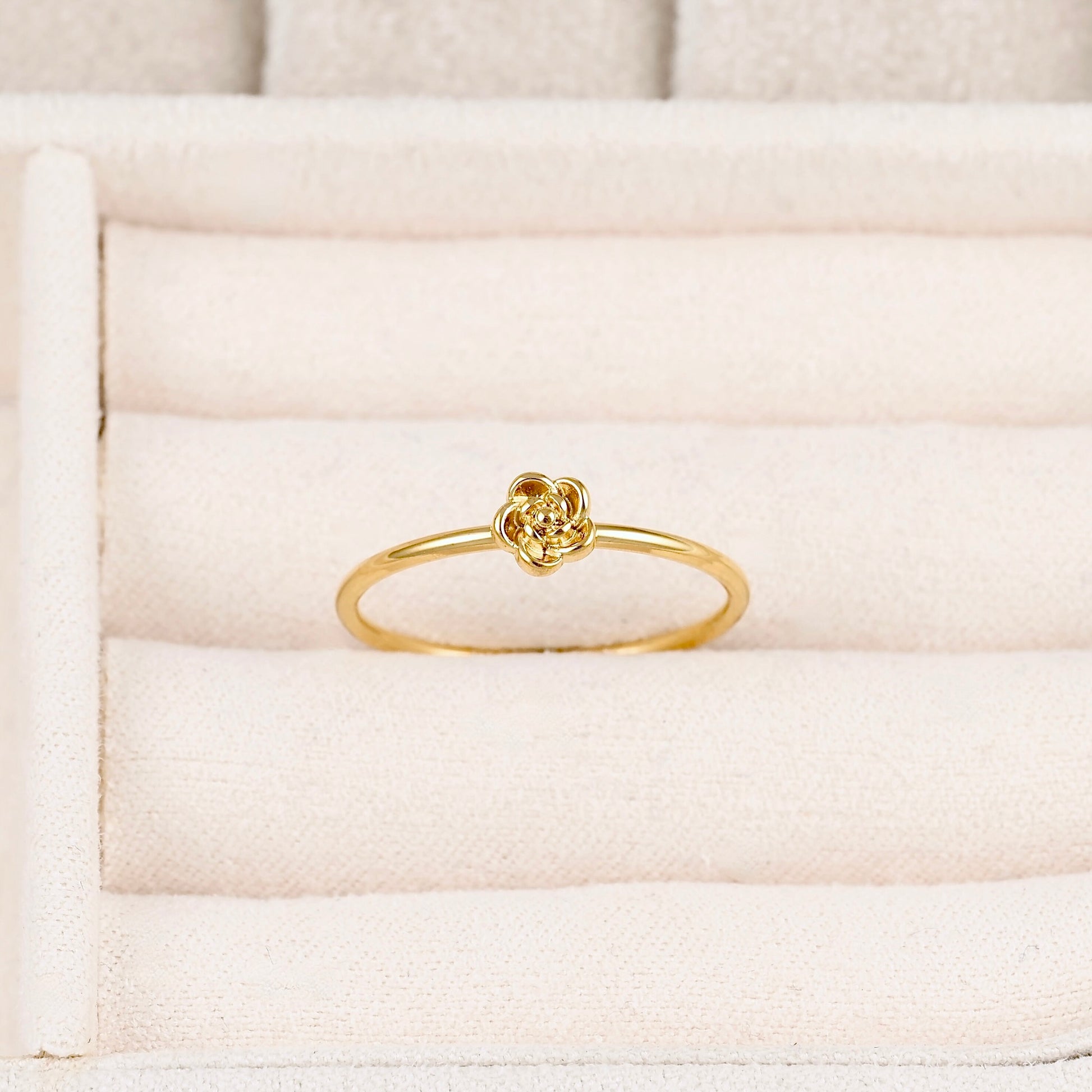 A delicate yellow gold ring featuring a small, intricate floral or knot-like top, displayed on a cream-colored textured ring box.