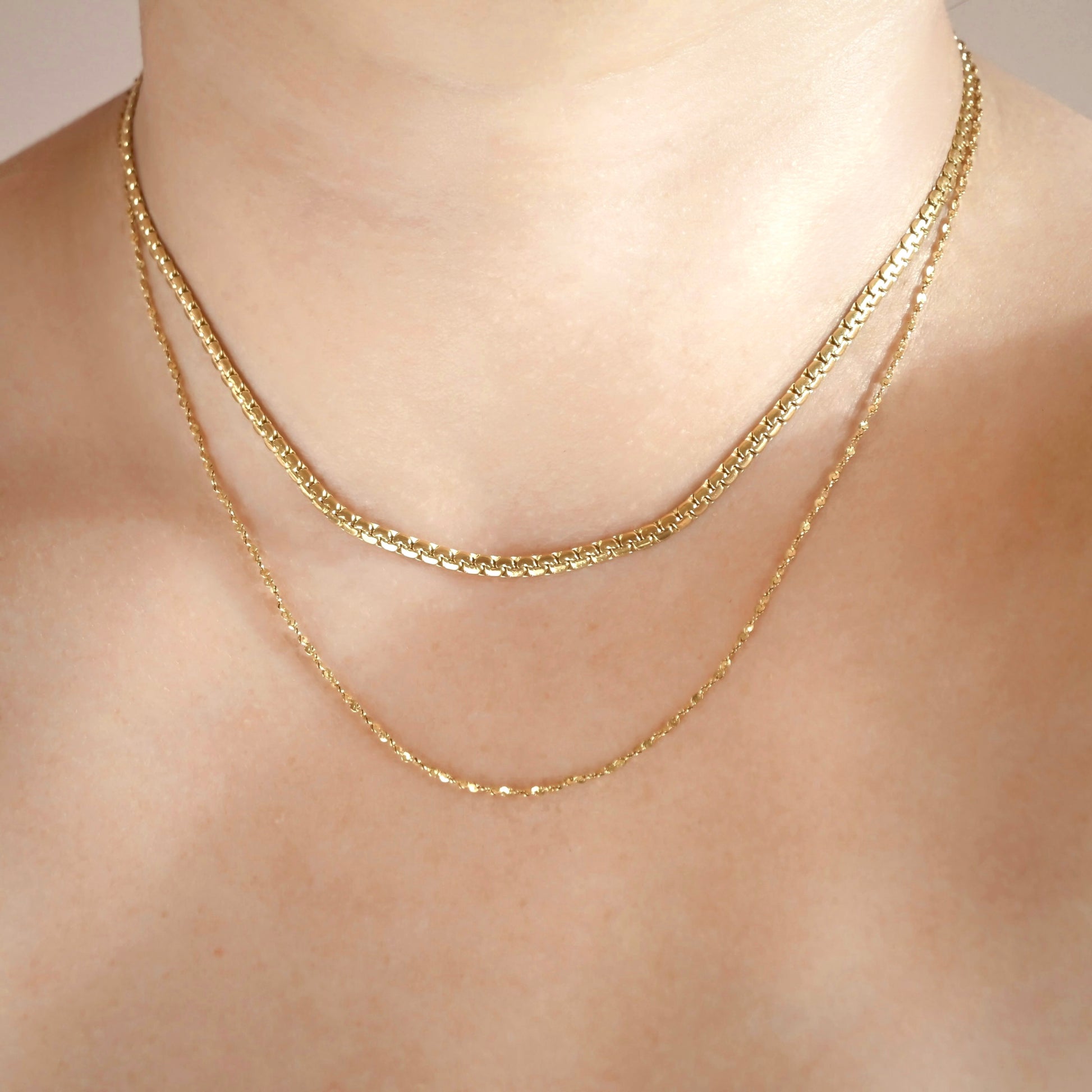 Two delicate gold chain necklaces layered on a model's neck, featuring a slightly thicker flat curb chain on top and a finer sparkle chain below.