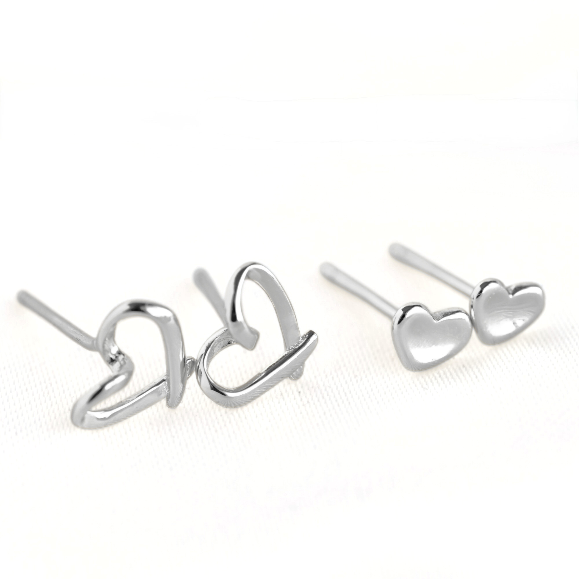 A pair of silver stud earrings, one in a heart  outline shape and one as small solid hearts, displayed on a clean white textured background.