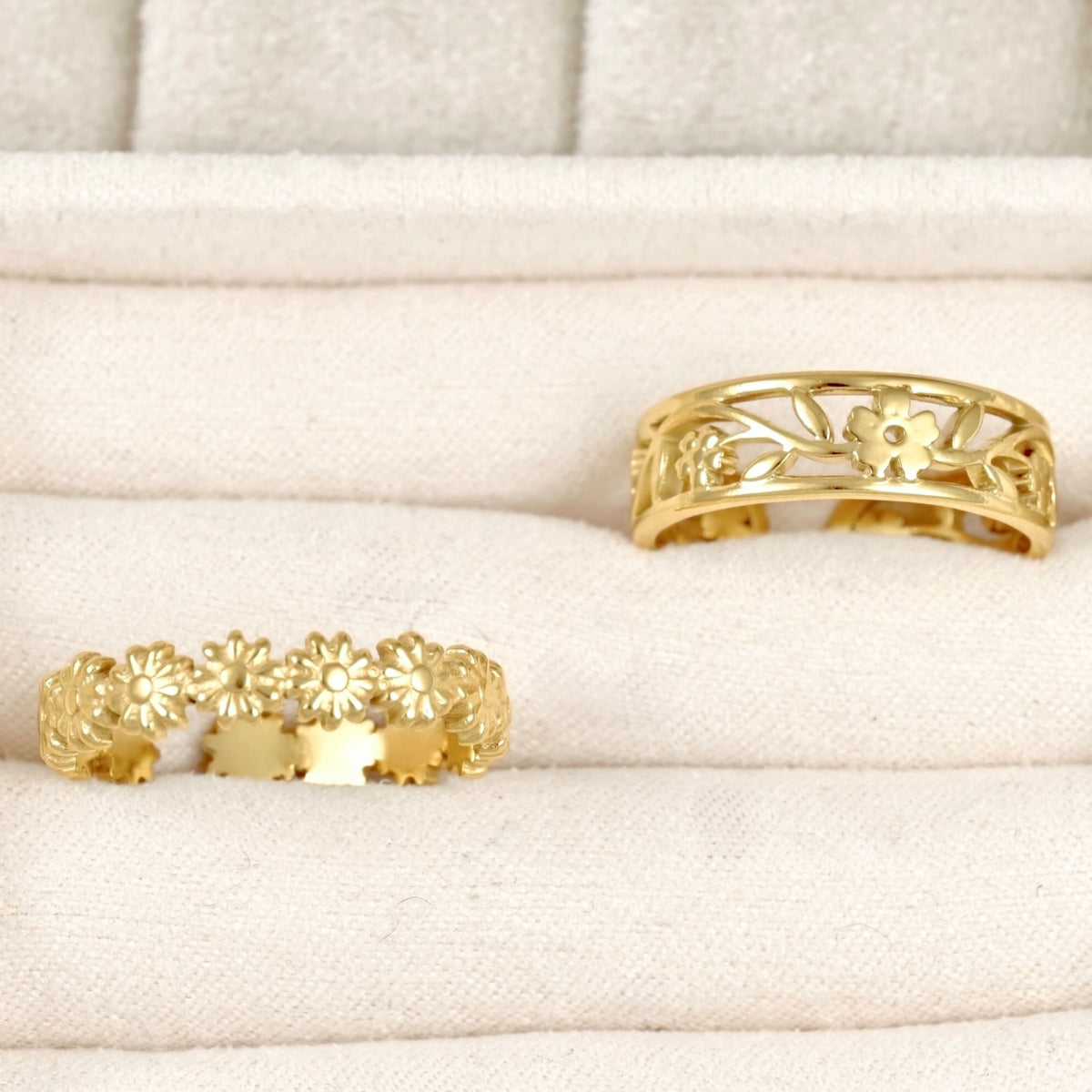 Two gold rings displayed on a cream-colored padded jewelry box. One ring features a continuous row of small flower motifs, while the other has an openwork floral and leaf pattern.