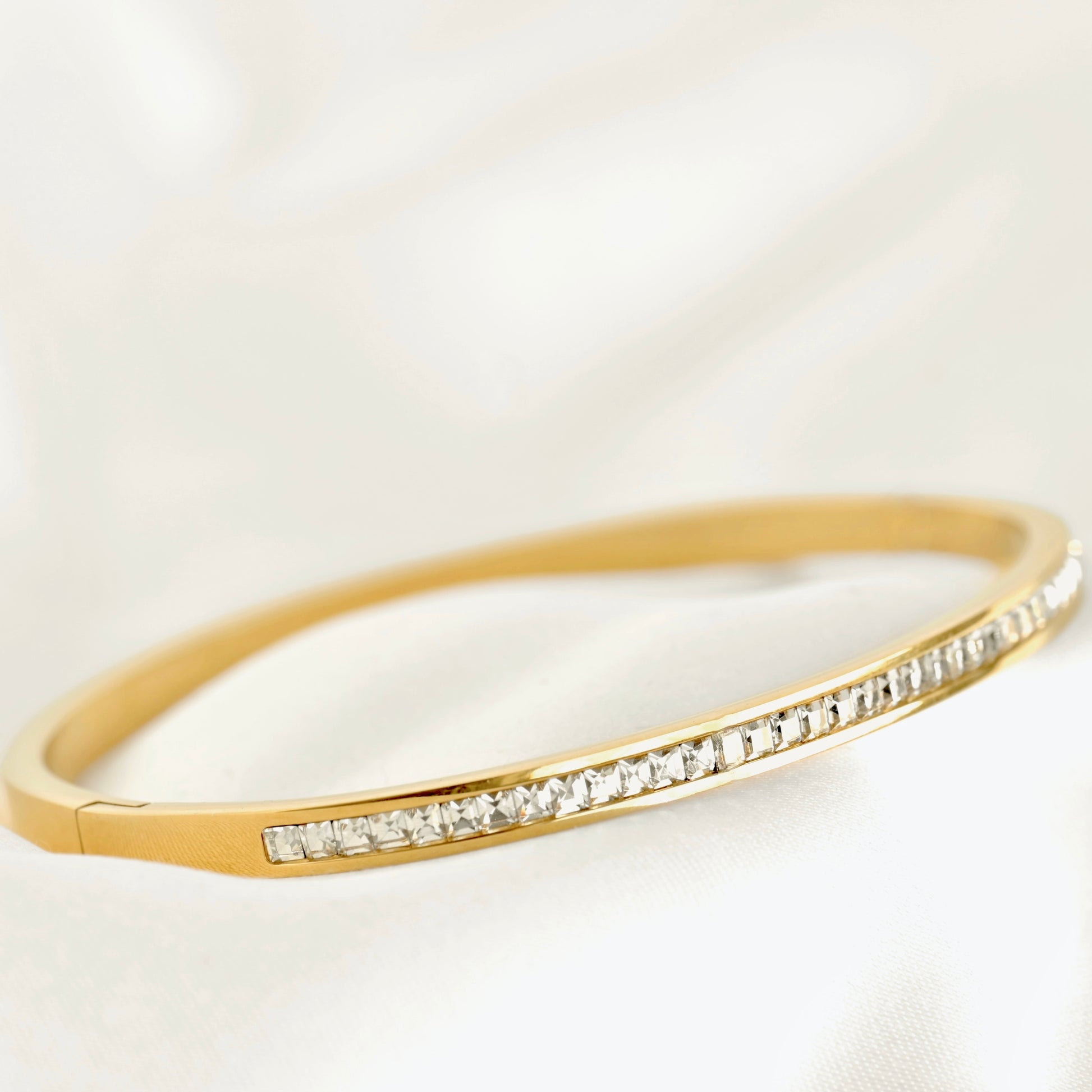 A gold hinged bangle bracelet with a channel-set row of clear stones along the front, displayed on a soft white fabric background.