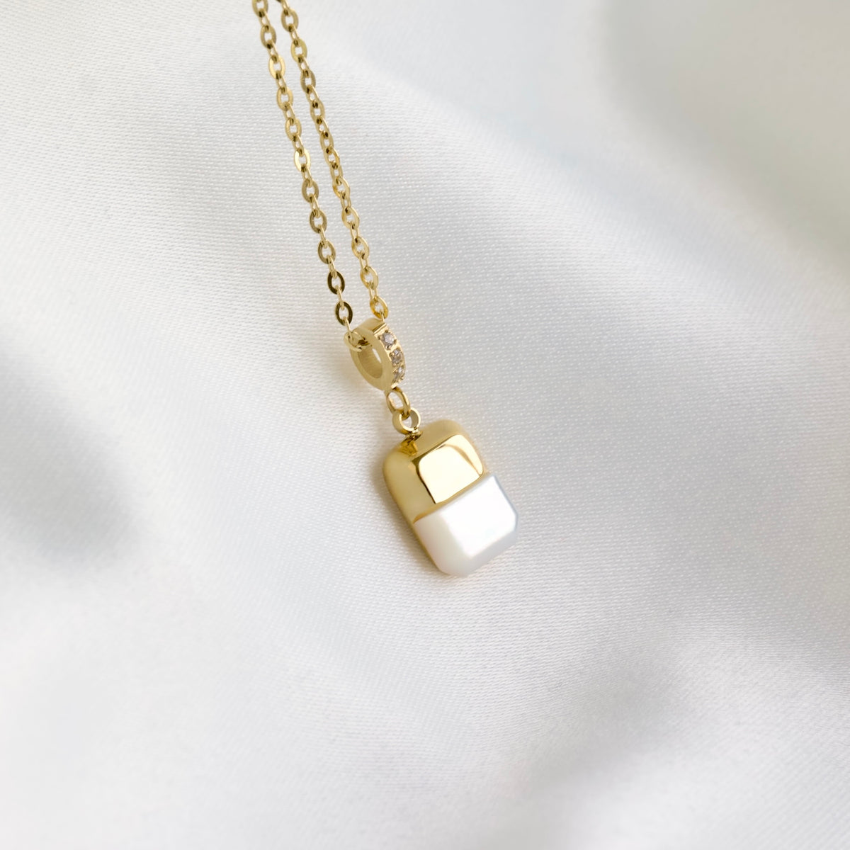 A gold chain necklace with a small capsule-shaped pendant, half gold and half white pearl, resting on a soft white fabric background.