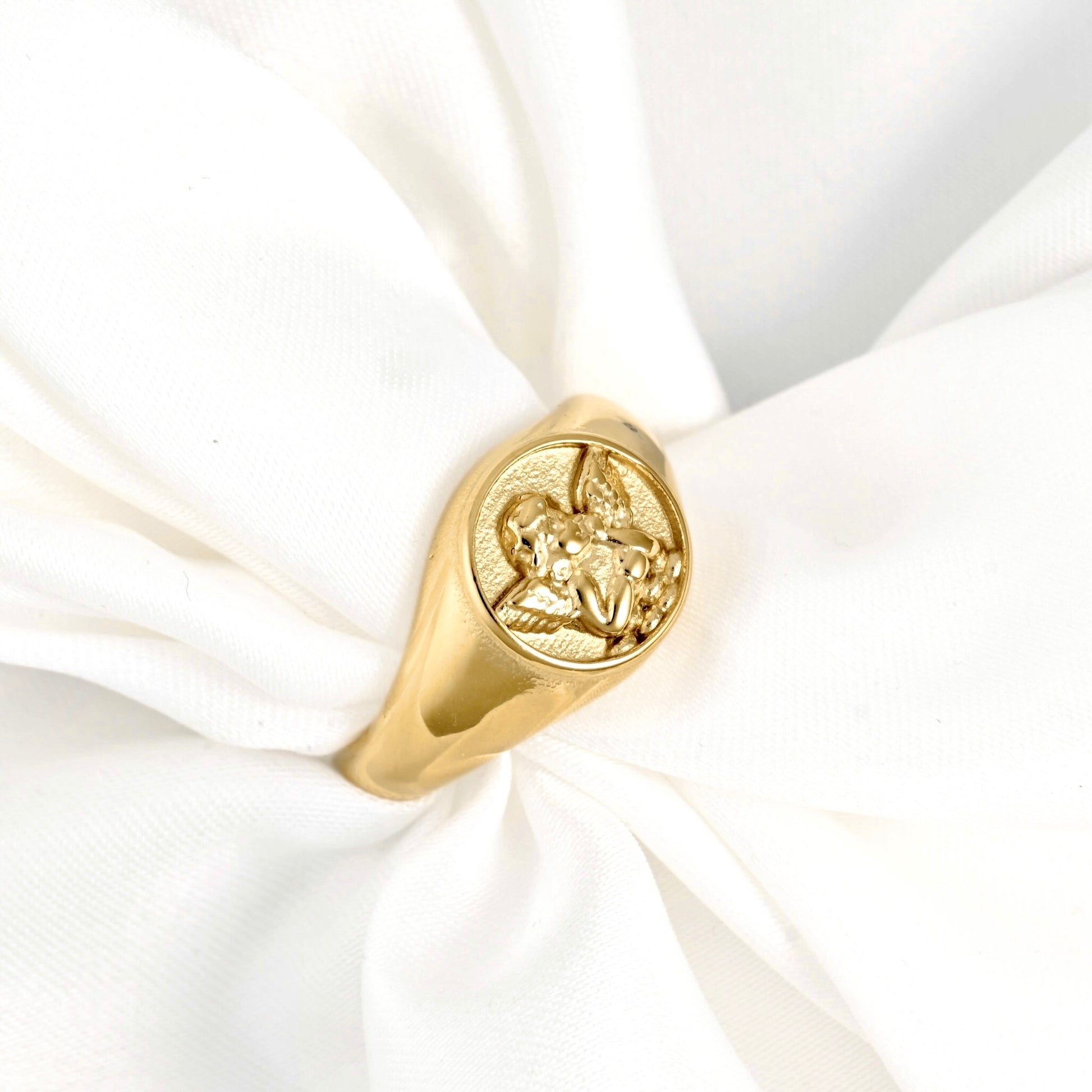 A gold signet ring featuring an embossed angel design, displayed on a soft, white fabric ring holder.