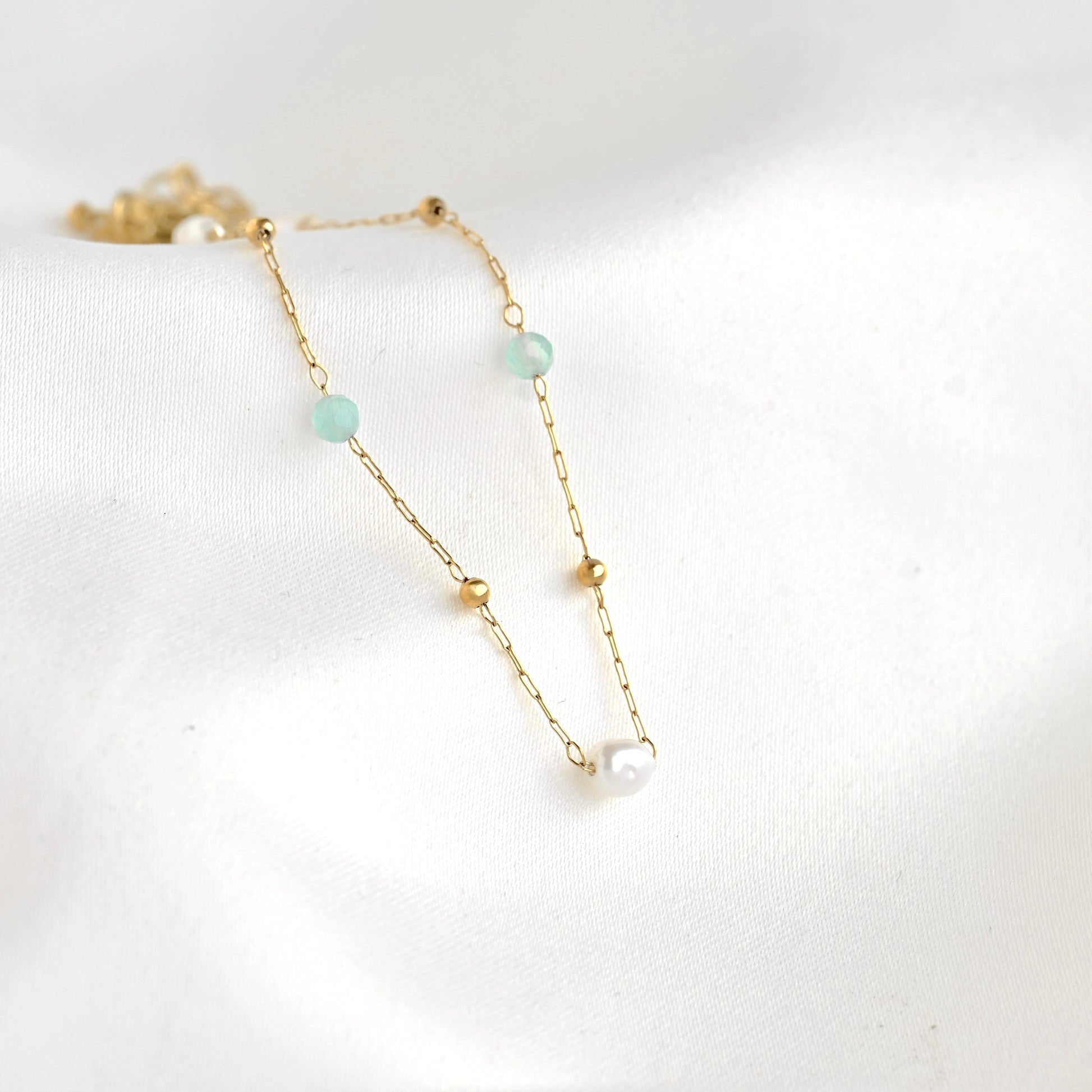 A delicate gold chain bracelet featuring light green round beads, small gold spacer beads, and a single white pearl connector, displayed on a soft white fabric background.
