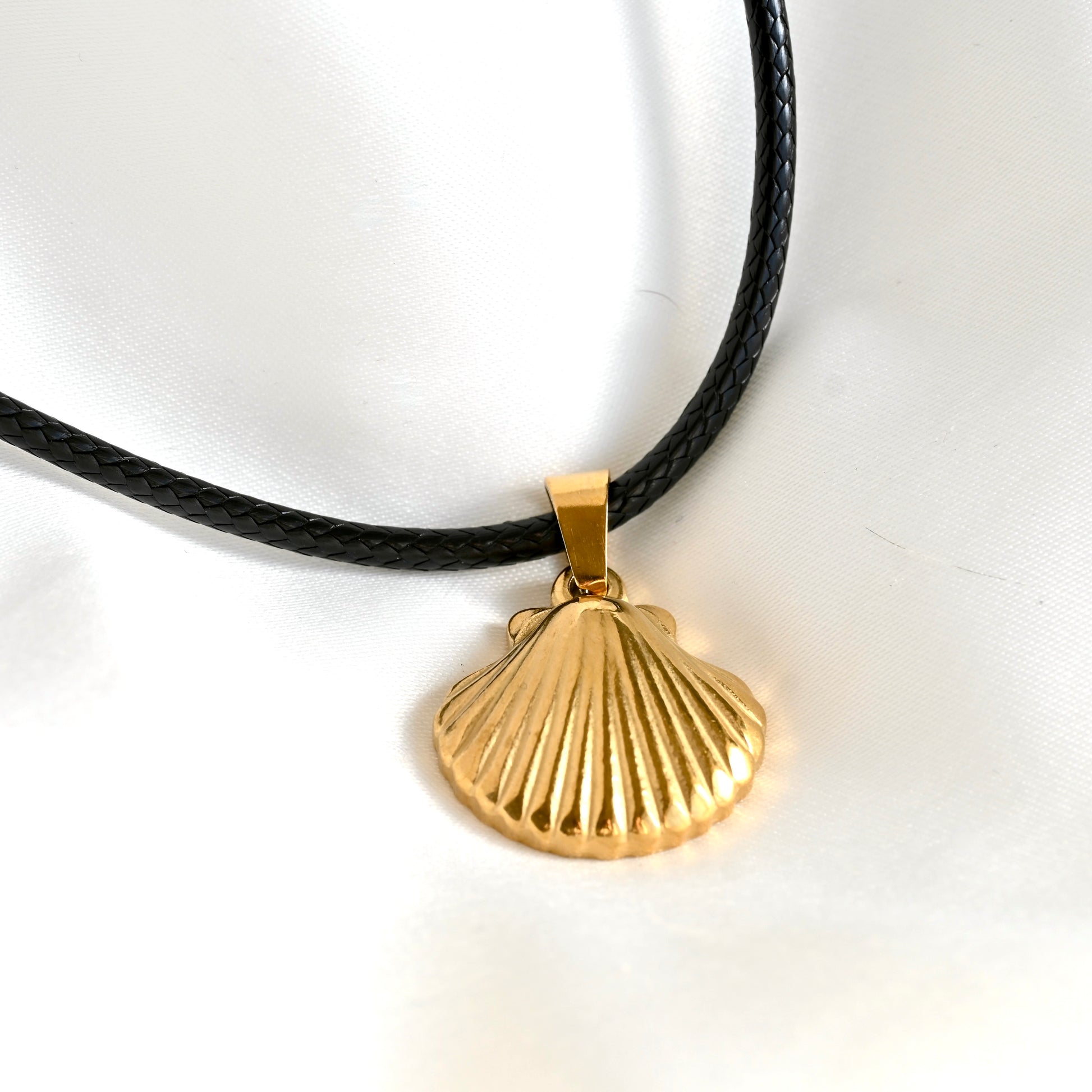 A gold scallop shell pendant necklace on a black braided waxed cord, photographed on a white fabric background.