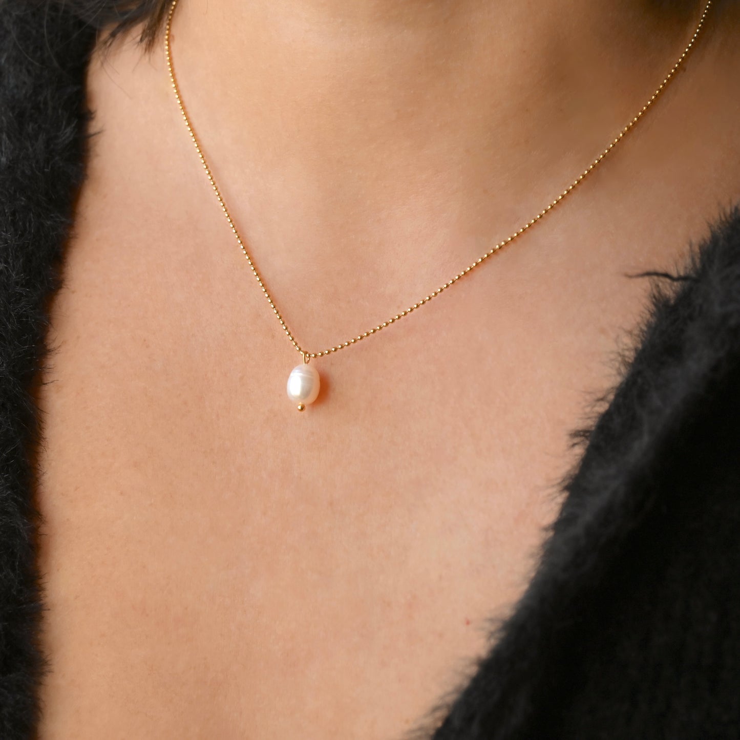 A close-up of a delicate gold chain necklace with a single teardrop-shaped white pearl pendant, worn on a model's neck.