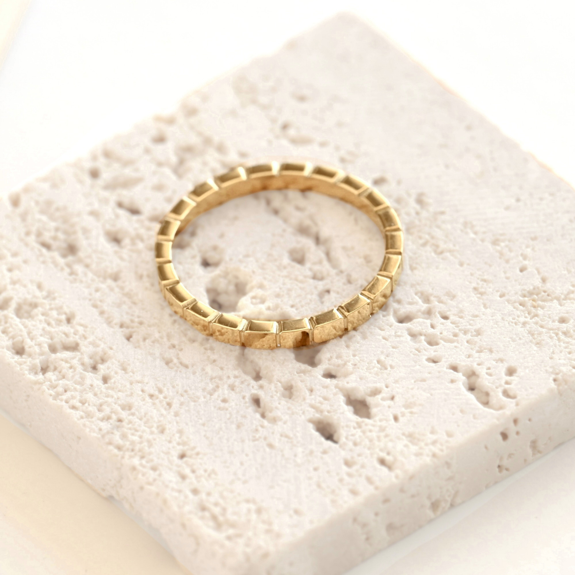 A gold ring with a textured, block-like segmented design, displayed on a square, light beige stone tile against a clean white background.