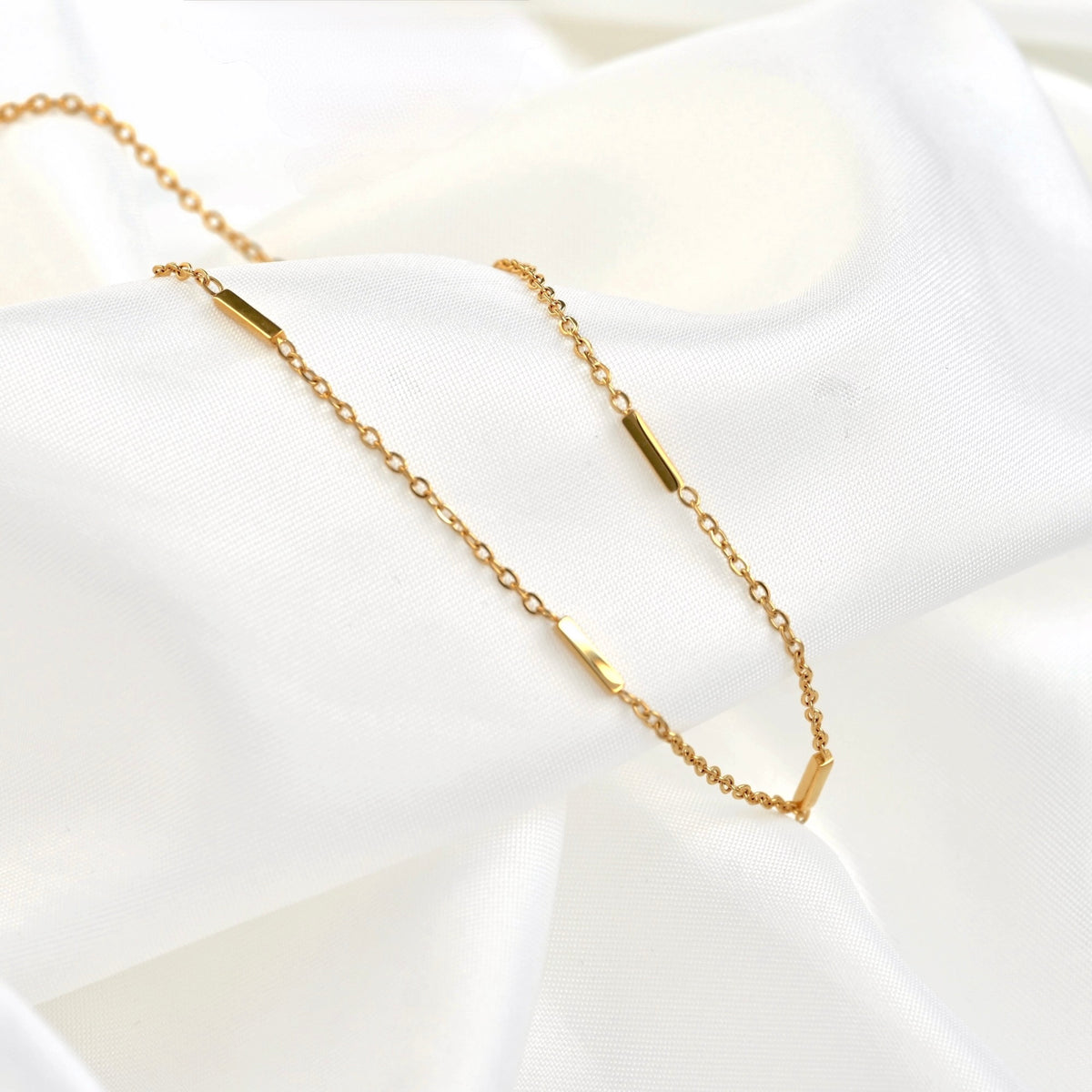 A delicate gold chain necklace with small polished gold bar charms, displayed on draped white fabric.