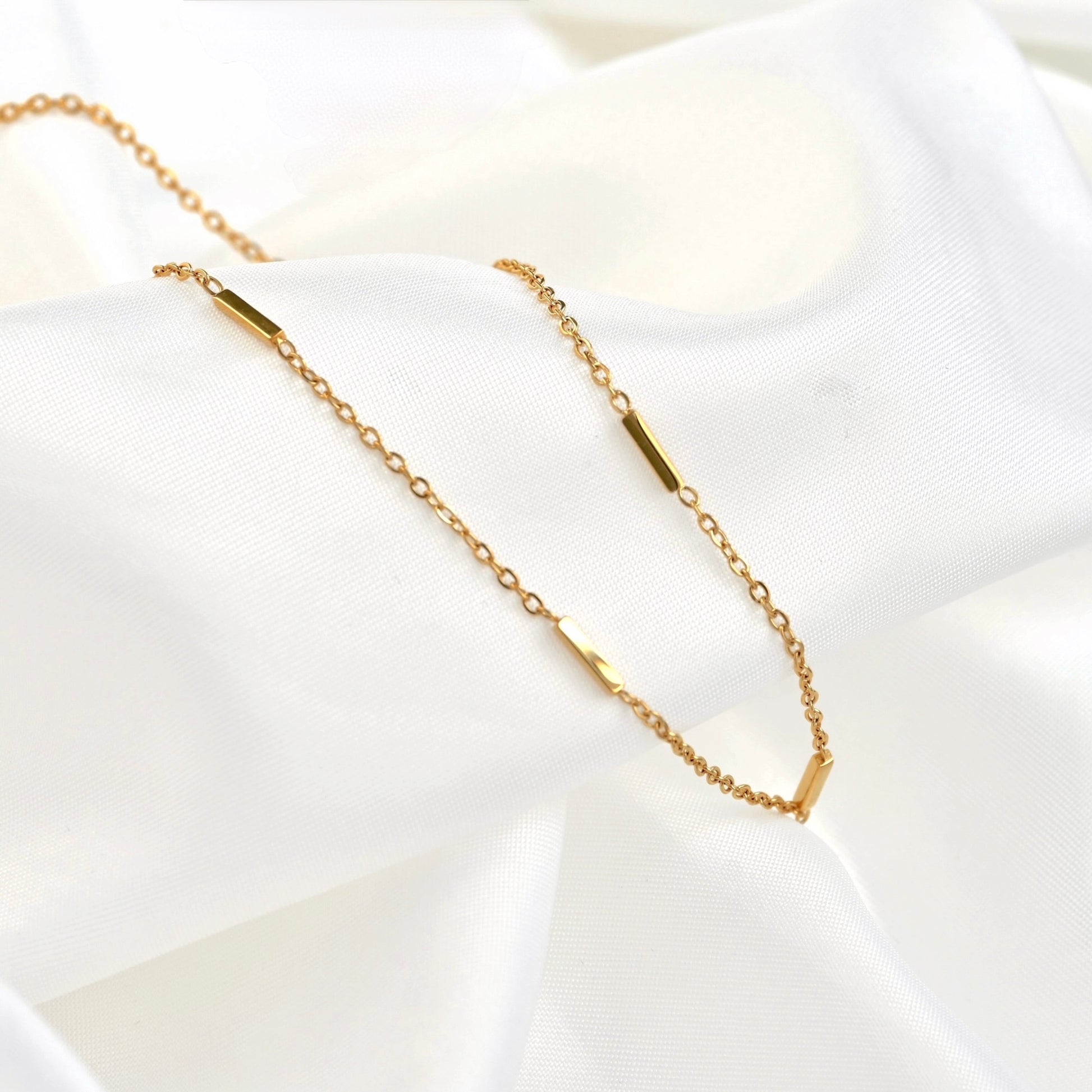 A delicate gold chain necklace with small polished gold bar charms, displayed on draped white fabric.
