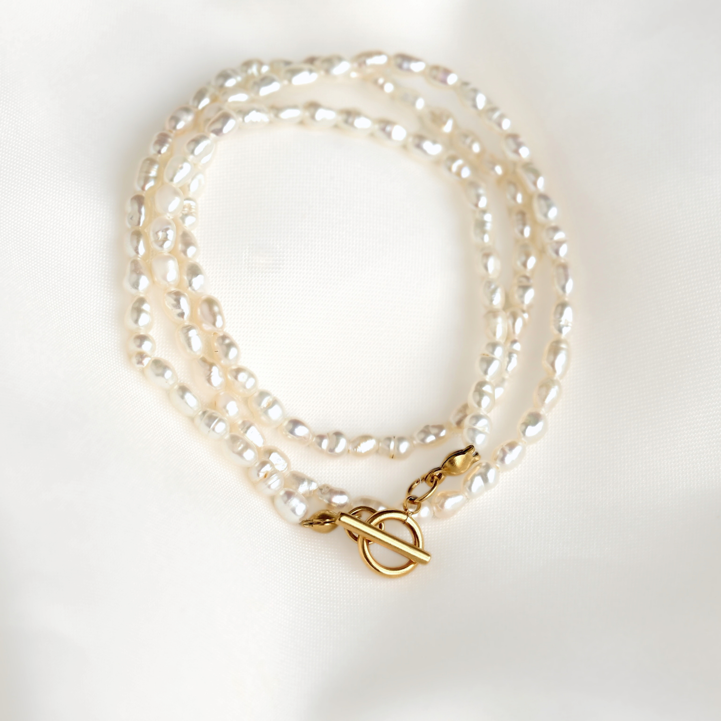 A thin rice pearl necklace with a gold toggle clasp, displayed on a soft white fabric background.