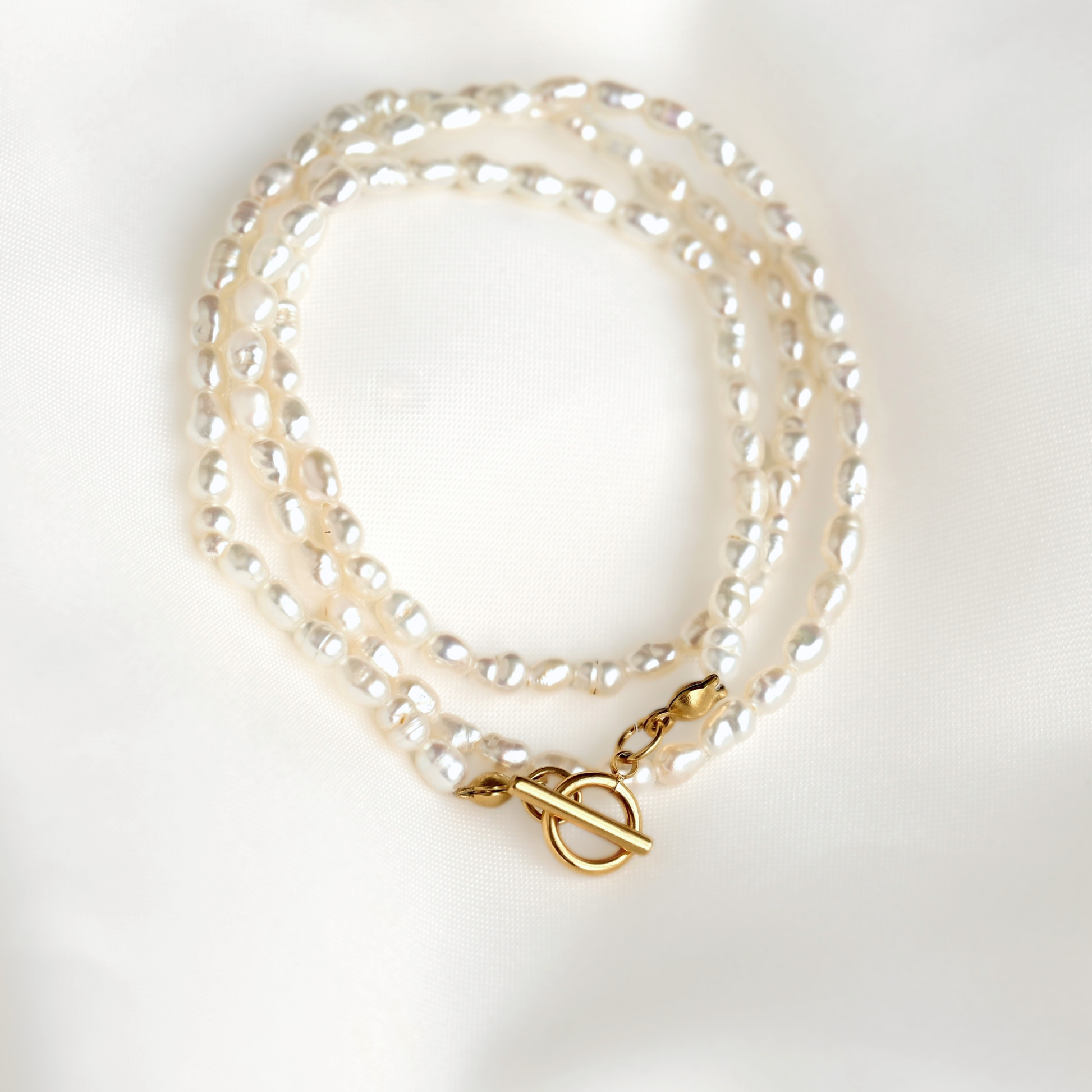 A thin rice pearl necklace with a gold toggle clasp, displayed on a soft white fabric background.