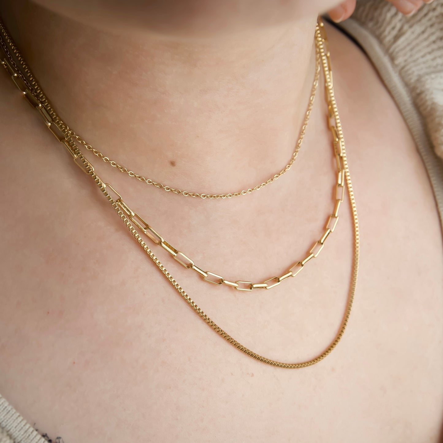 Close-up of a person wearing a gold multi-strand chain necklace featuring cable, paperclip and box chains on a neutral background