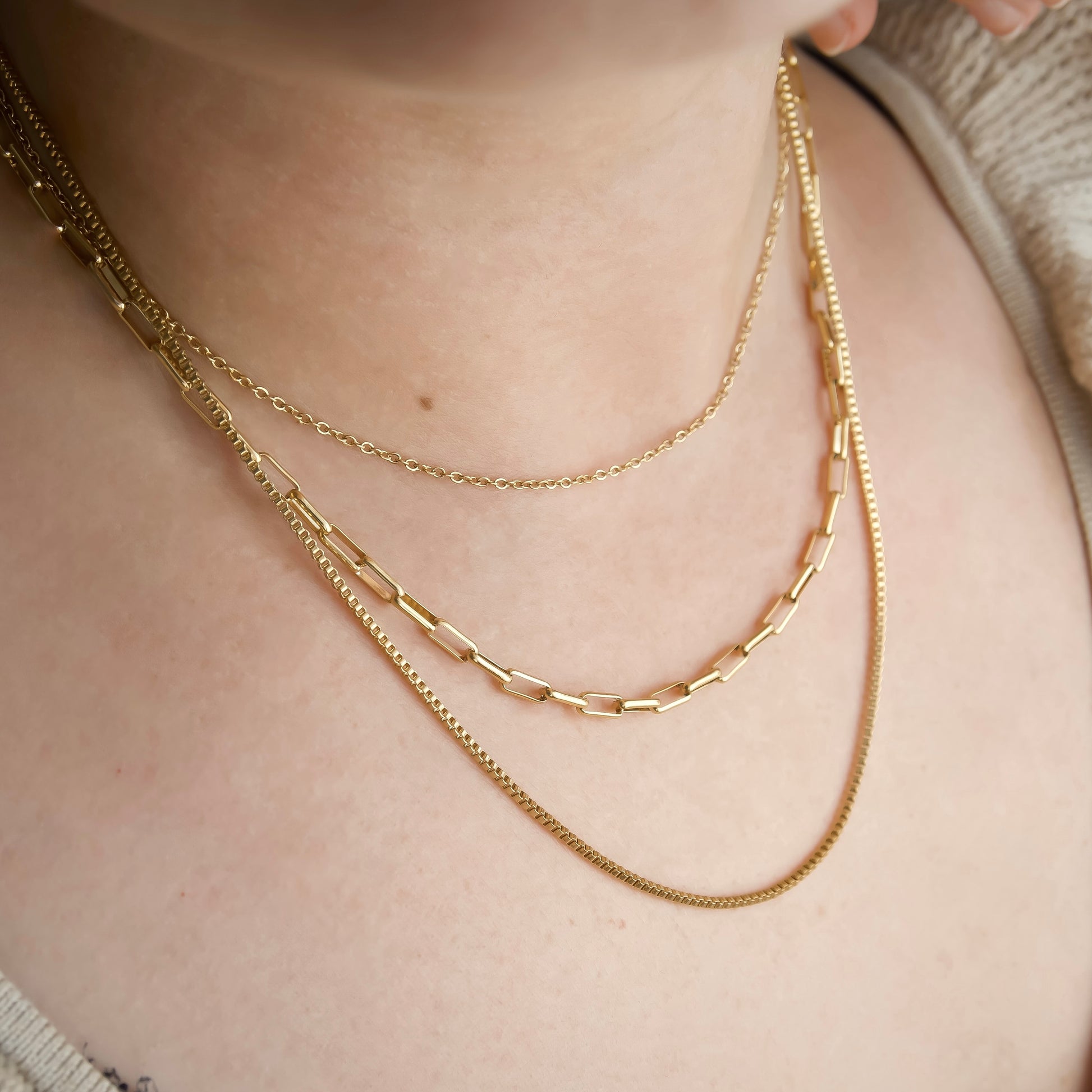 Close-up of a person wearing a gold multi-strand chain necklace featuring cable, paperclip and box chains on a neutral background