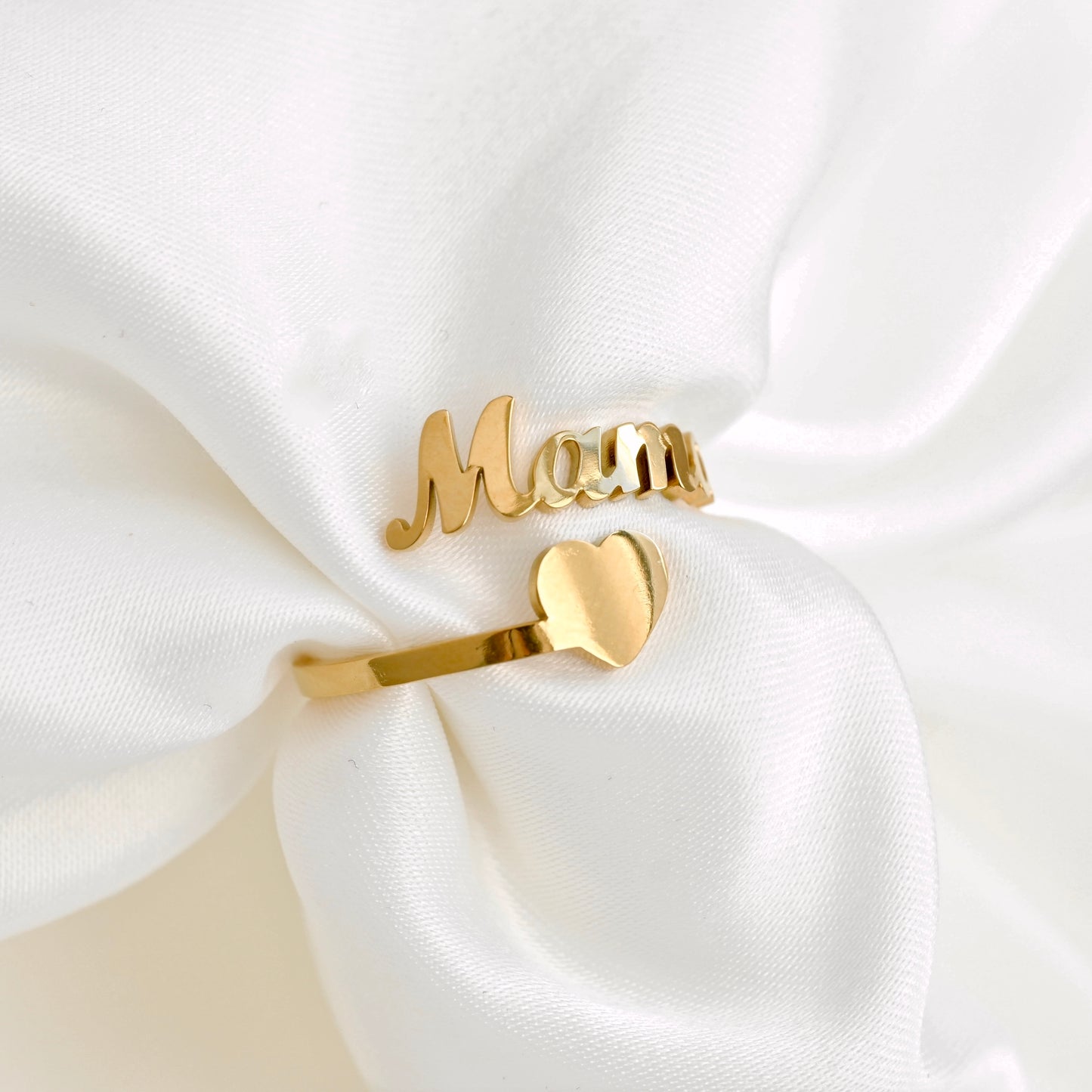 A gold bypass ring displayed on a white satin fabric background, featuring the word 'Mama' in cursive script on one end, an a simple heart shape on the other end.