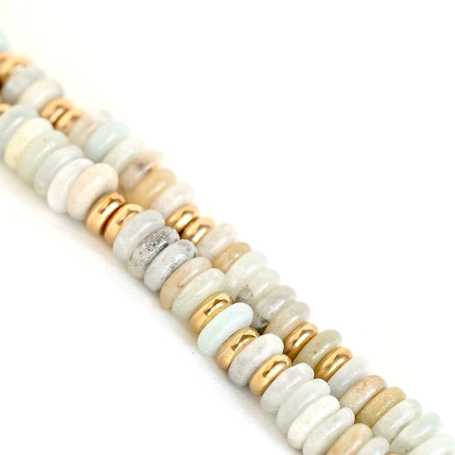 A close-up diagonal view of a beaded strand featuring polished round stone beads in soft gray, cream, and light beige, interspersed with small metallic gold spacer beads, set against a clean white background.