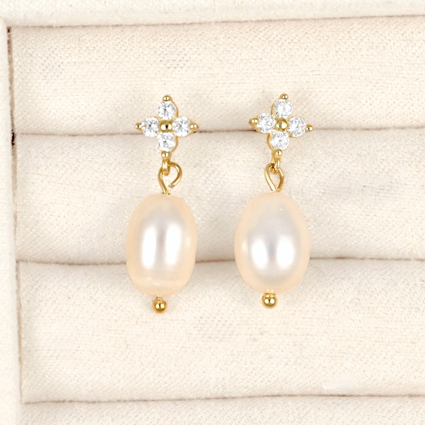 A pair of gold drop earrings with oval cream-colored freshwater pearls and clear crystal floral studs, displayed on a cream-colored textured jewelry box.