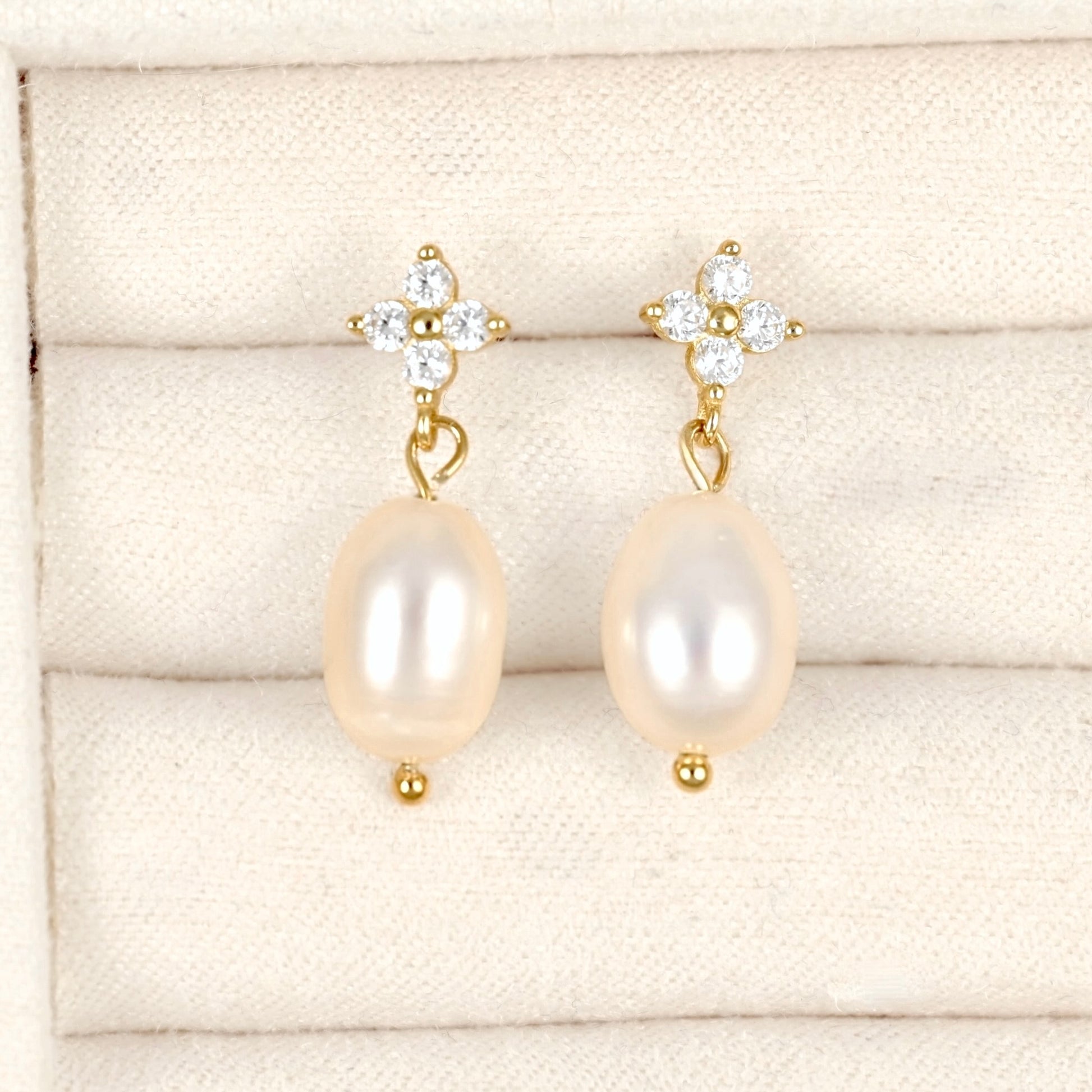 A pair of gold drop earrings with oval cream-colored freshwater pearls and clear crystal floral studs, displayed on a cream-colored textured jewelry box.