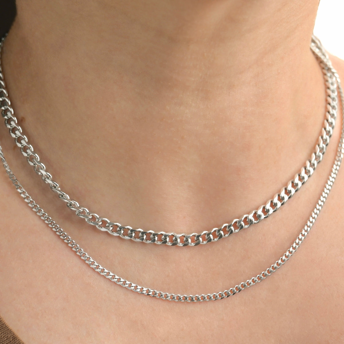 A close-up of a person's neck wearing two layered silver-tone chain necklaces, including one chunky and one finer curb chain.