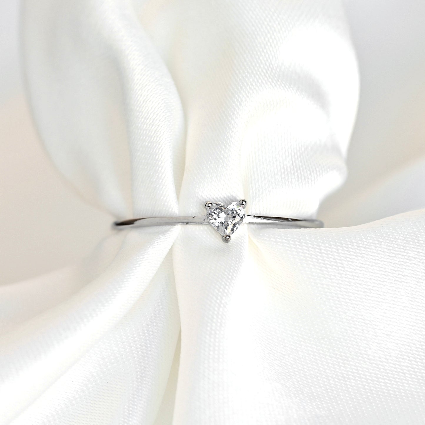 A dainty silver diamond ring with a small triangular clear gemstone, displayed on a soft, draped white satin fabric.