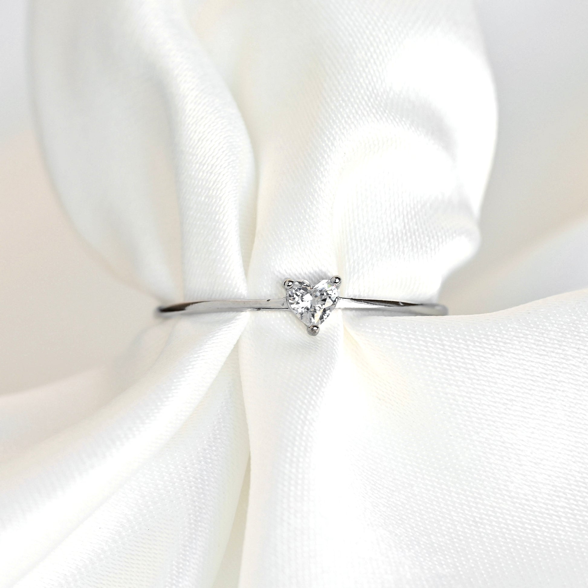 A dainty silver diamond ring with a small triangular clear gemstone, displayed on a soft, draped white satin fabric.