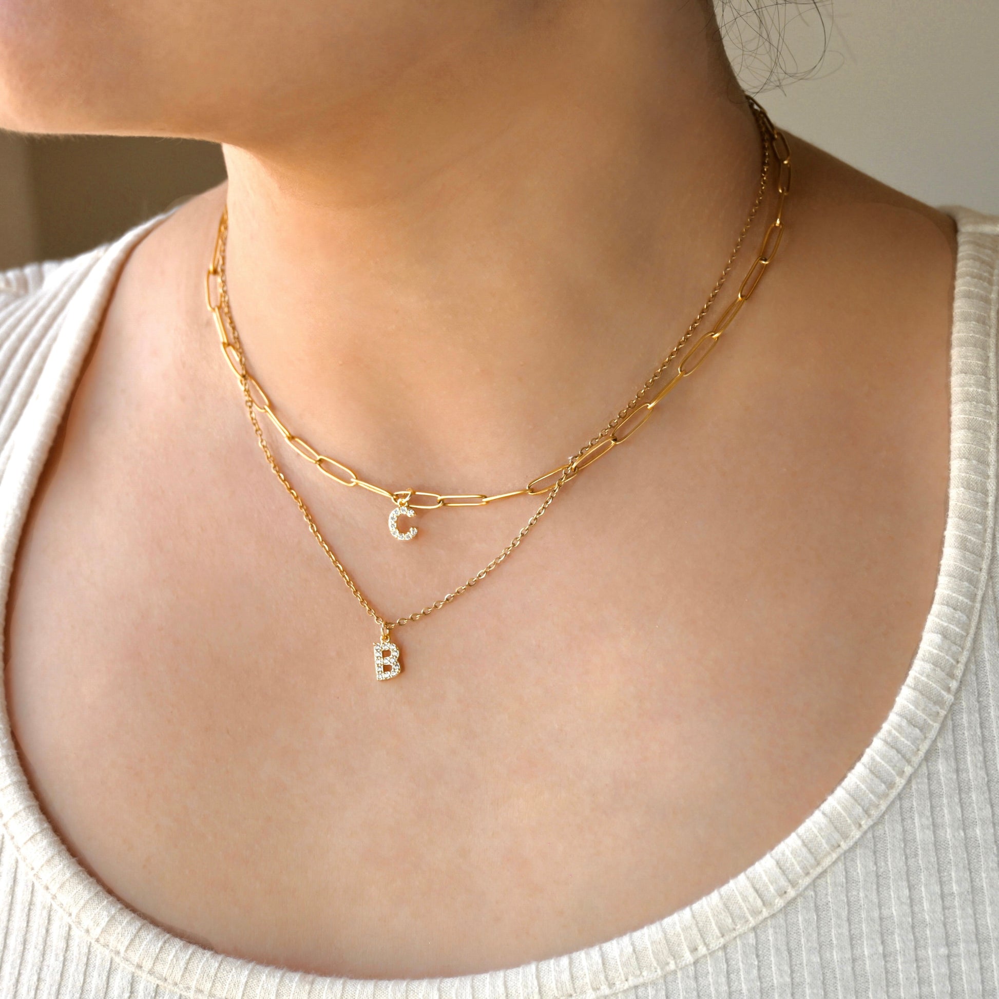 Two gold necklaces layered, one shorter paperclip chain with letter 'C', and one longer cable chain with letter 'B' on a person's neck
