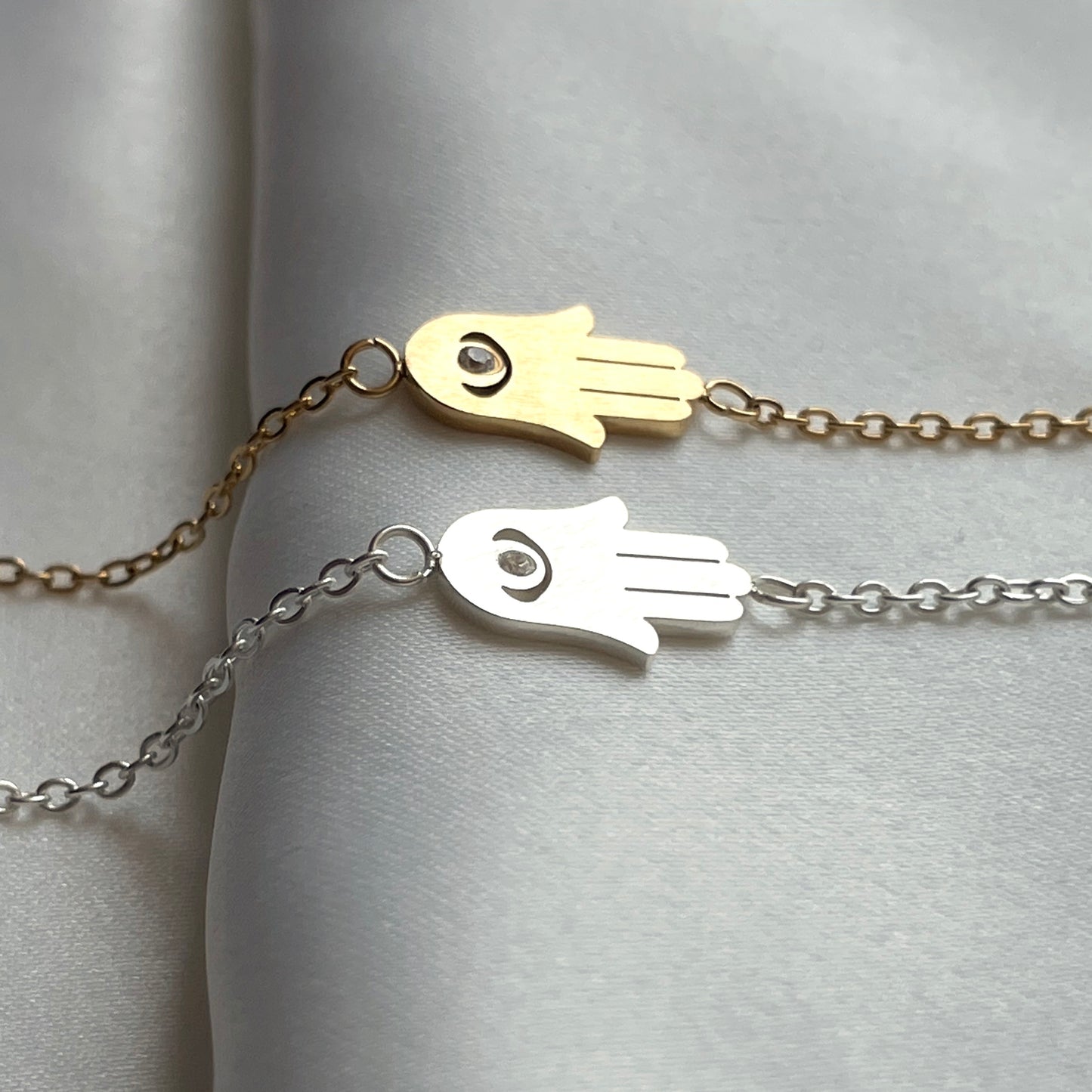Two Hamsa hand necklaces, one gold and one silver, on a light gray background.