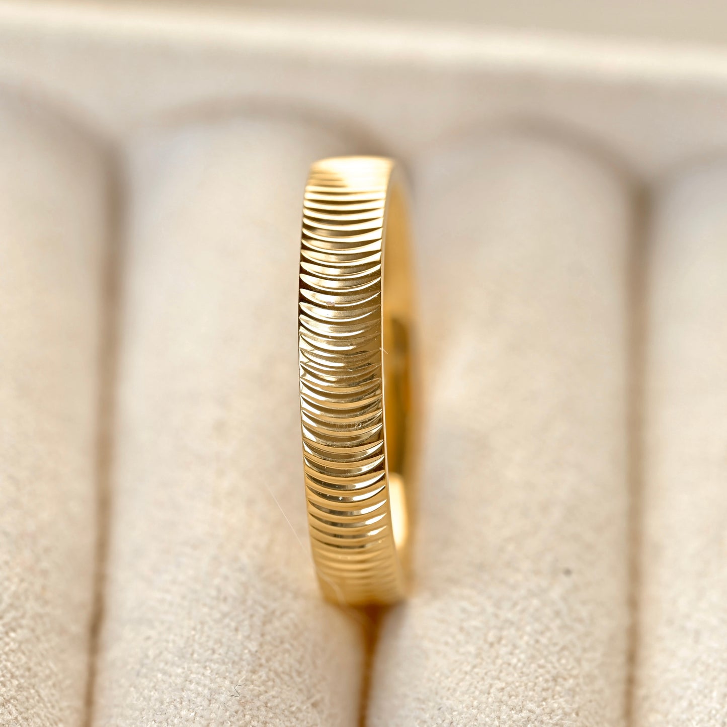 A close-up of a textured gold ring with horizontal ridges, displayed on a beige velvet ring box.