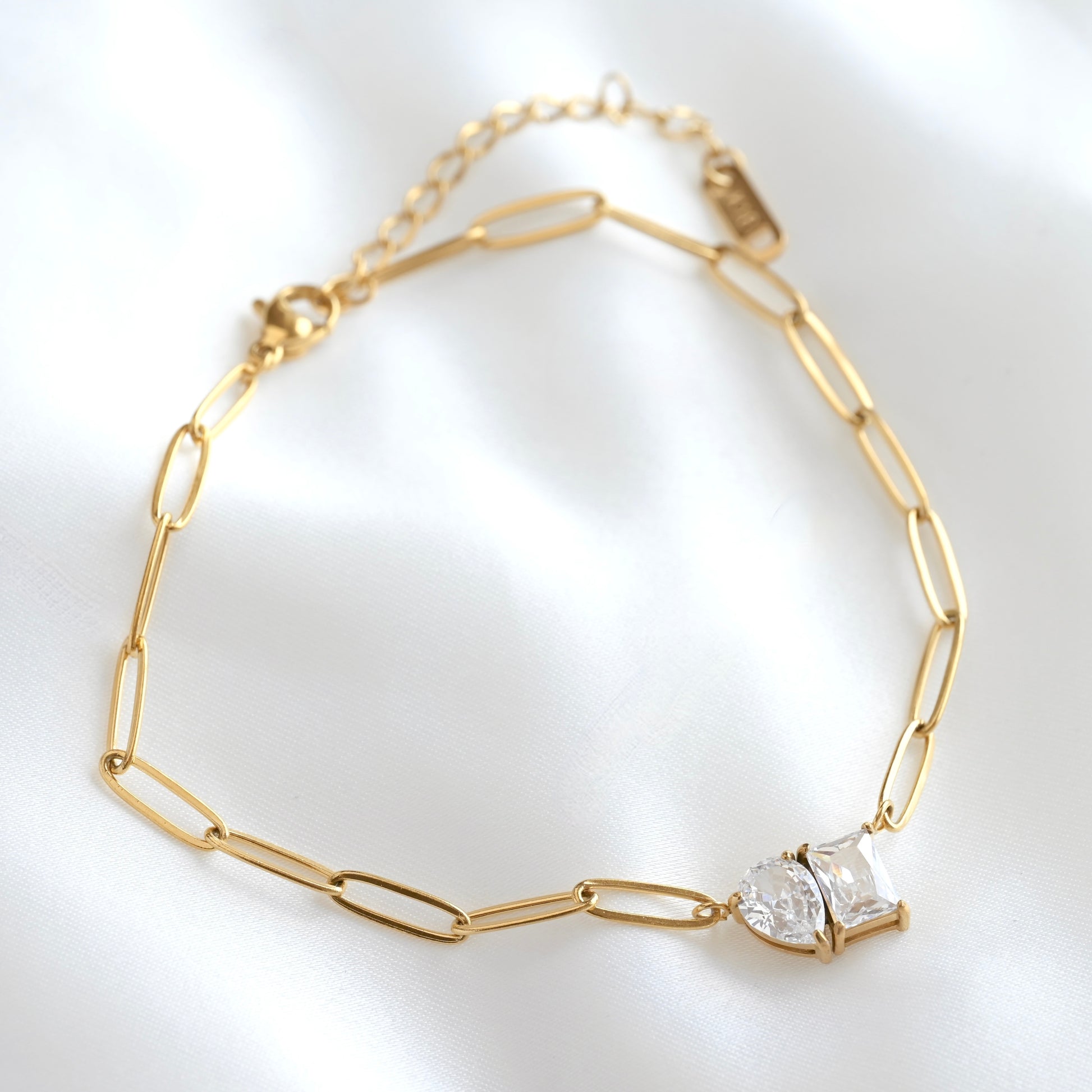 A gold paperclip chain necklace with a toi et moi stone pendant, displayed on a soft white satin fabric background.