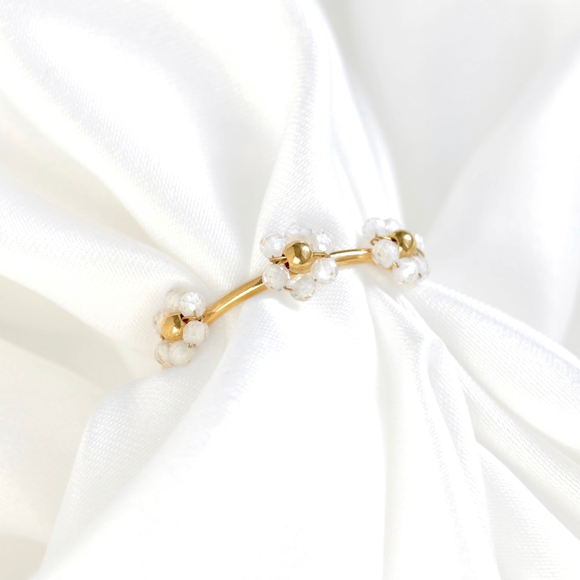 A delicate gold flower ring featuring small white pearl flower clusters, displayed on a soft, draped white satin fabric.