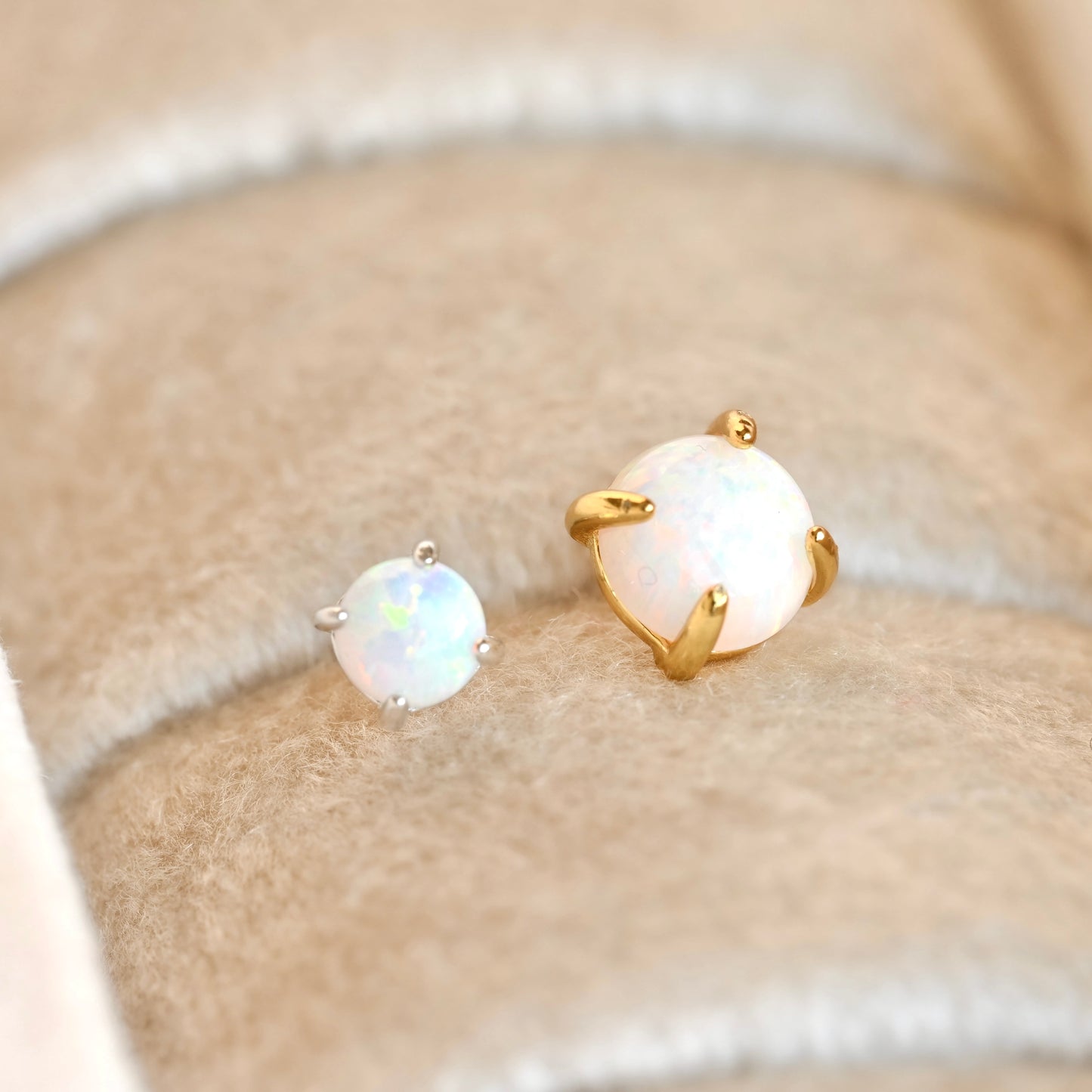 A pair of stud earrings with round white opal gemstones in gold and silver settings, displayed on a beige felt jewelry ring cushion.